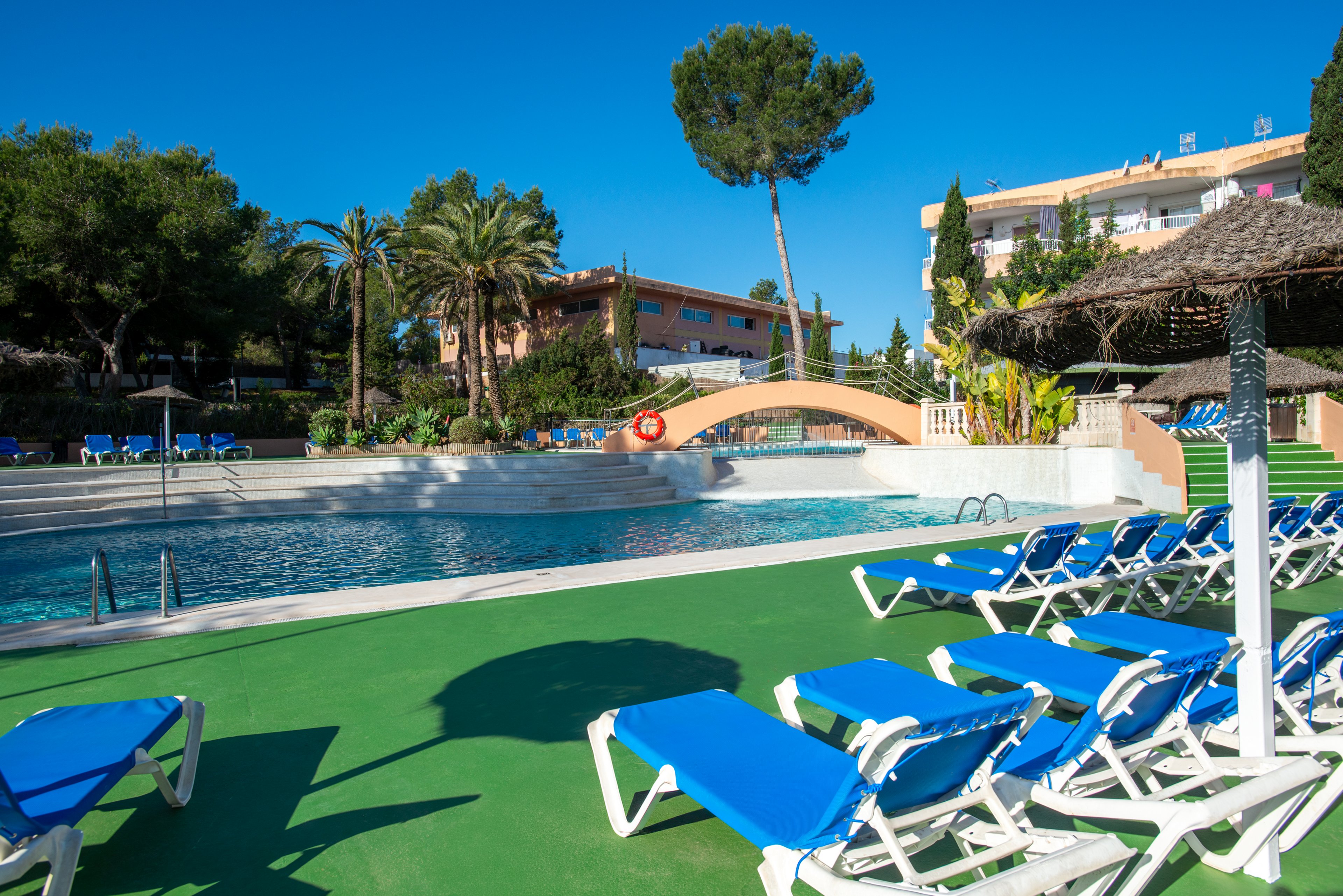 An outdoor resort swimming pool is surrounded by rows of empty blue sun loungers on a bright, sunny day. A small arched bridge crosses part of the pool, with a red lifebuoy hanging on its railing. Palm trees and lush greenery frame the scene, while low-rise buildings are visible in the background. The clear blue sky and strong shadows emphasize the warm, vacation atmosphere.