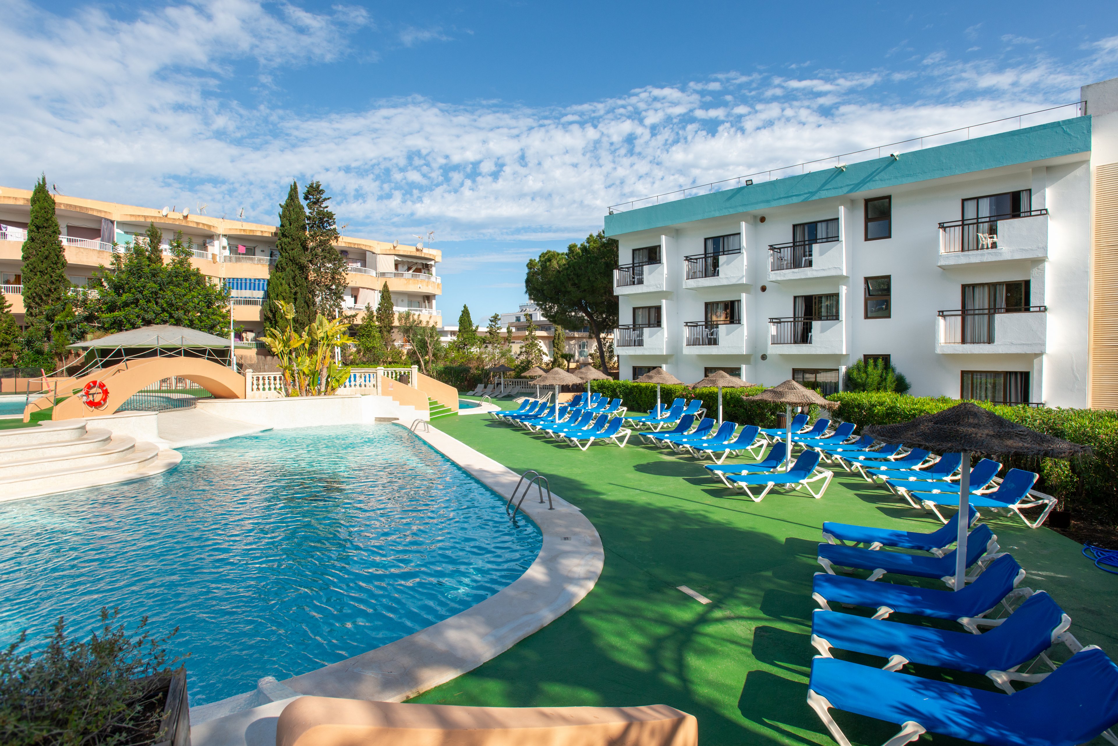 An outdoor swimming pool is surrounded by numerous blue lounge chairs at a sunny resort. A white multi-story hotel building with balconies overlooks the pool area. Green artificial turf and landscaped trees add color and contrast to the scene. The clear blue sky and bright light create a relaxed vacation atmosphere.