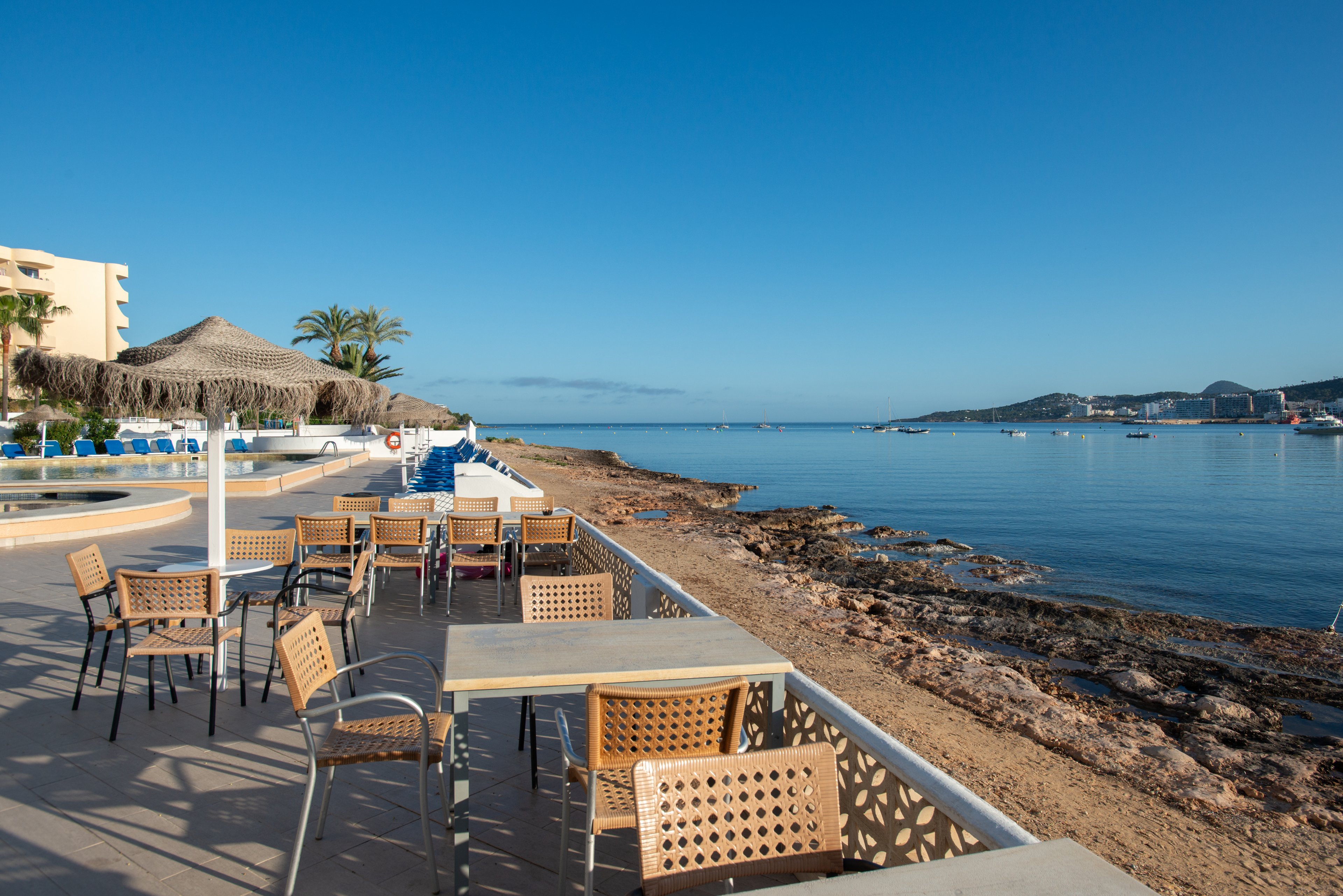 An outdoor resort terrace with empty tables and chairs overlooks a calm coastal bay. The seating area is set on a paved patio beside a rocky shoreline and clear blue water. Thatched umbrellas, palm trees, and sun loungers suggest a relaxing holiday setting. The sky is clear and bright, creating a peaceful, sunny atmosphere along the waterfront.