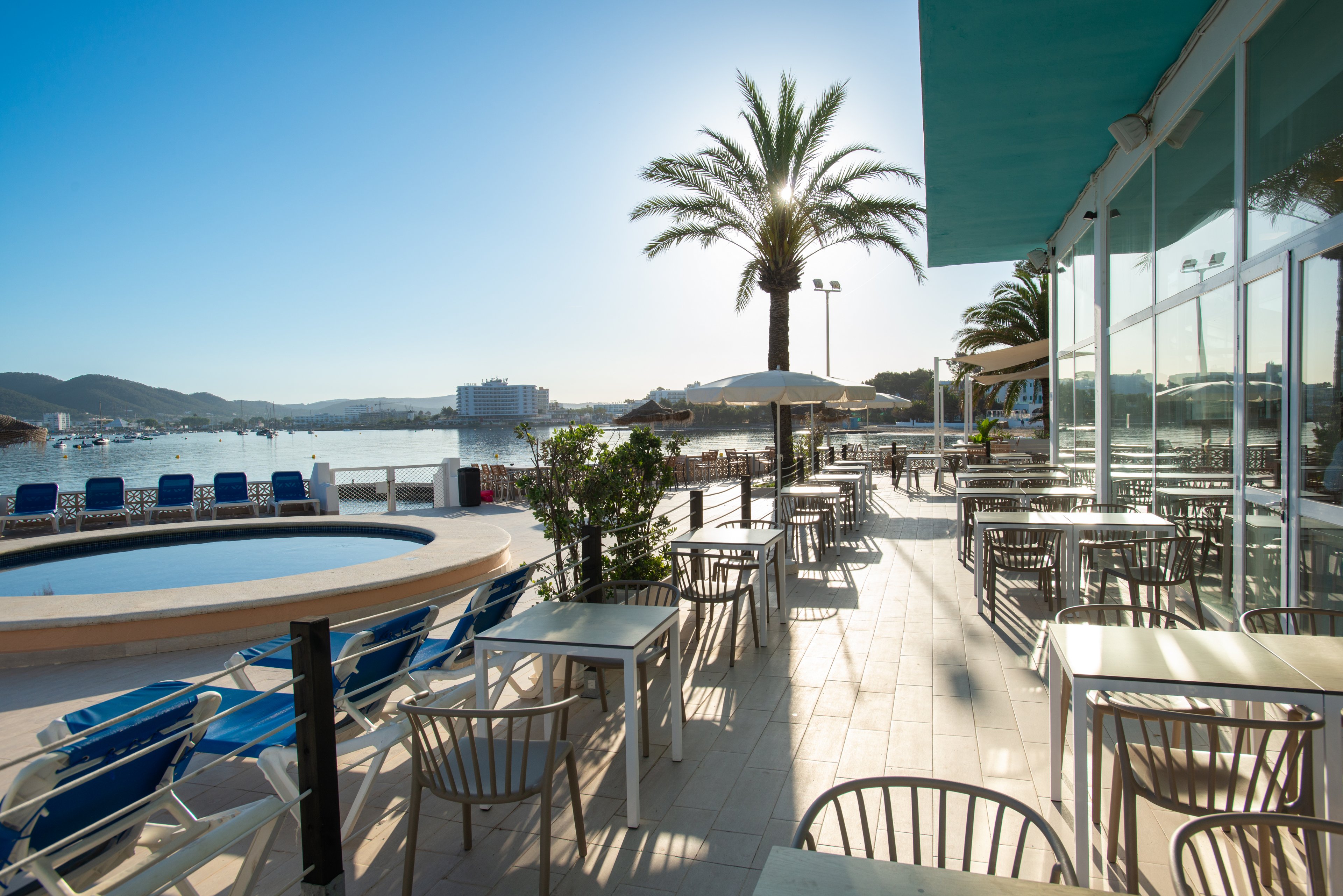 An empty outdoor terrace at a seaside resort is bathed in bright morning sunlight. Metal chairs and square tables line the tiled patio beside a small circular pool. Palm trees and potted plants add a tropical feel to the waterfront setting. In the distance, calm water, hills, and low-rise buildings complete the tranquil coastal scene.