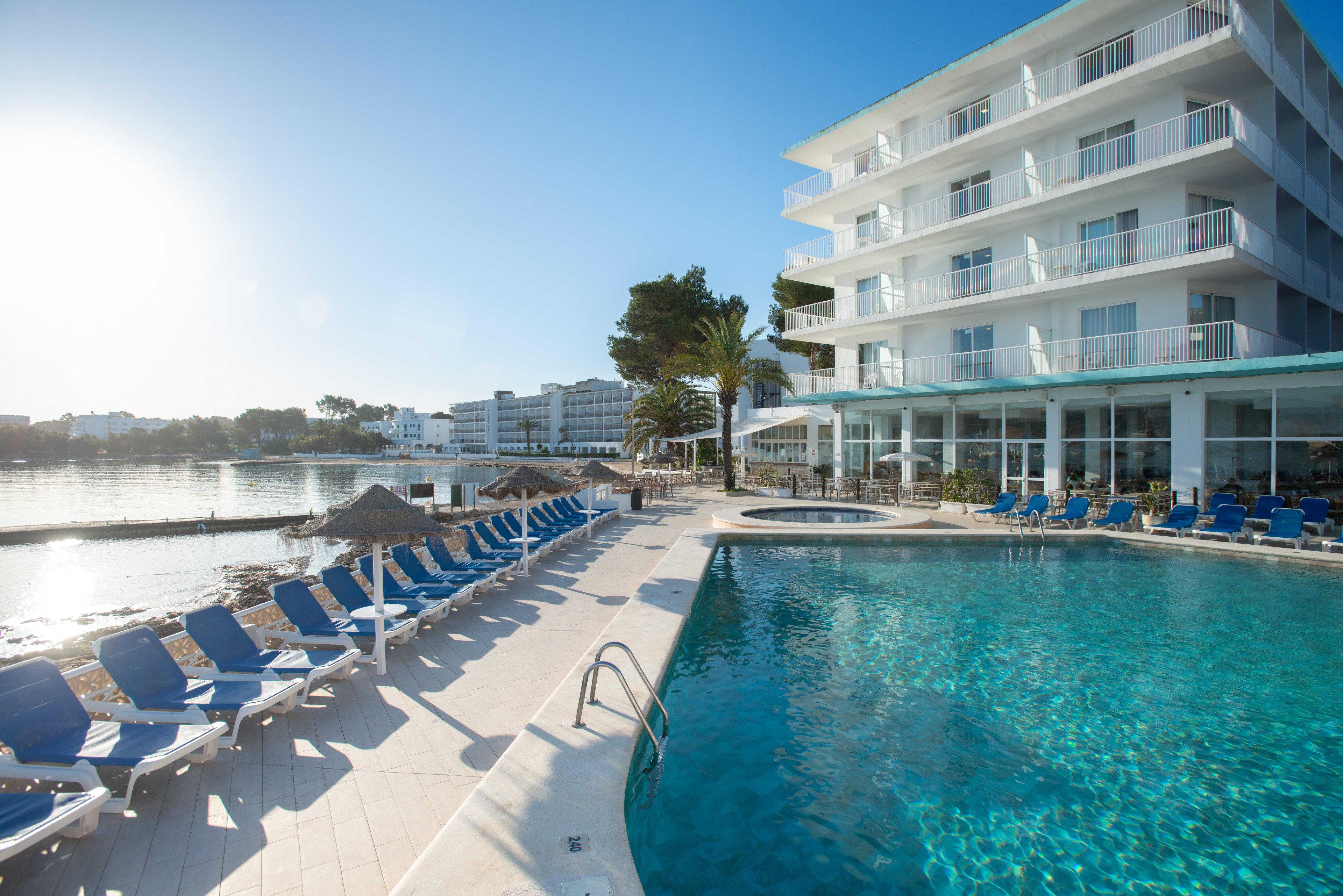 A contemporary multi-story hotel overlooks a large outdoor swimming pool filled with clear blue water. Numerous blue lounge chairs line the pool deck, facing the water and the nearby bay. The scene is set on a sunny day with a bright sky, calm water, and a few trees adding greenery. Glass windows on the ground floor suggest a restaurant or lobby area with views of the pool and waterfront.