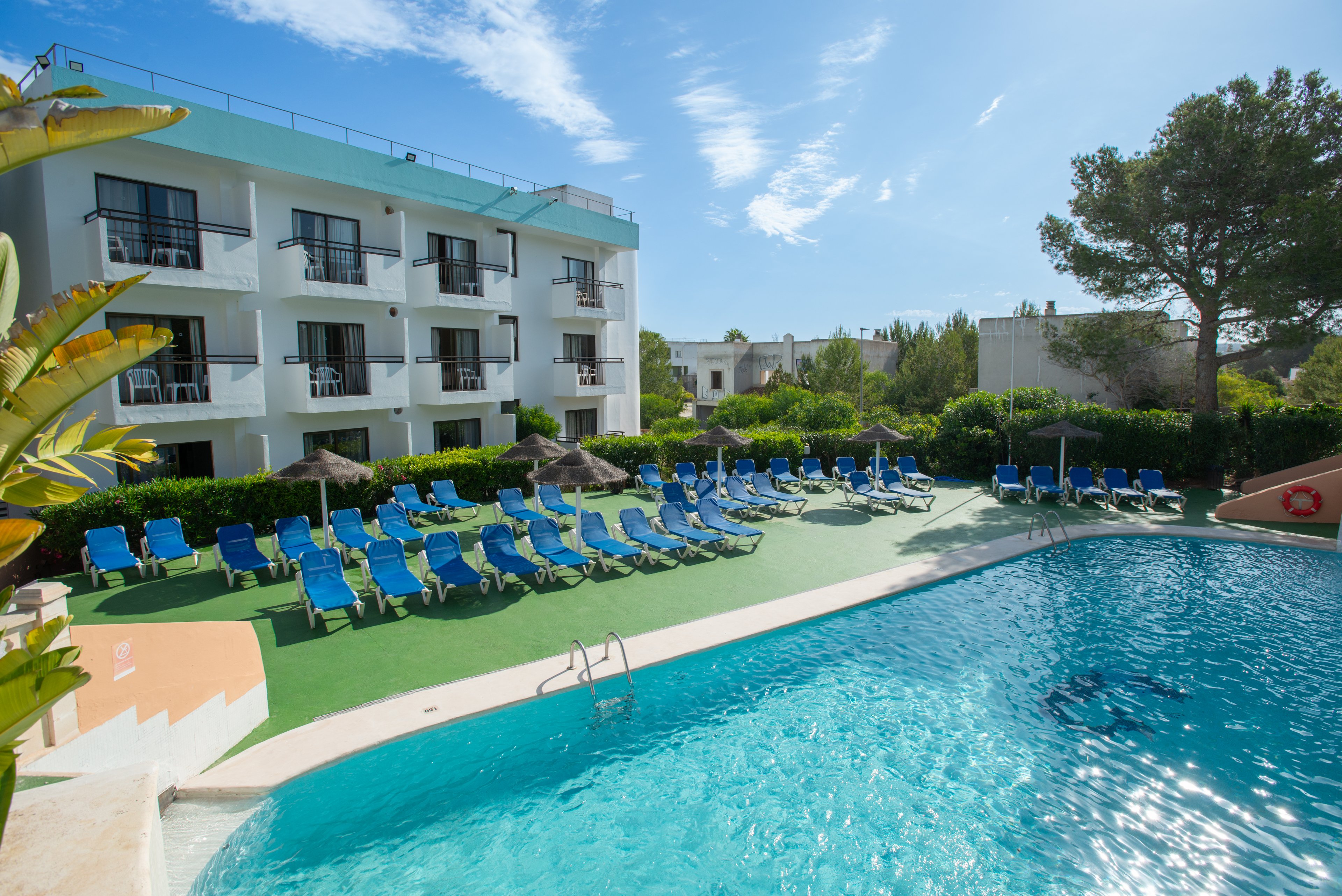 A bright outdoor swimming pool sits in front of a modern three-story hotel building. Numerous blue sun loungers are neatly arranged on a green poolside deck, all currently unoccupied. Lush greenery and trees surround the area, creating a secluded resort atmosphere. The clear blue sky and calm water suggest a warm, relaxing vacation setting.
