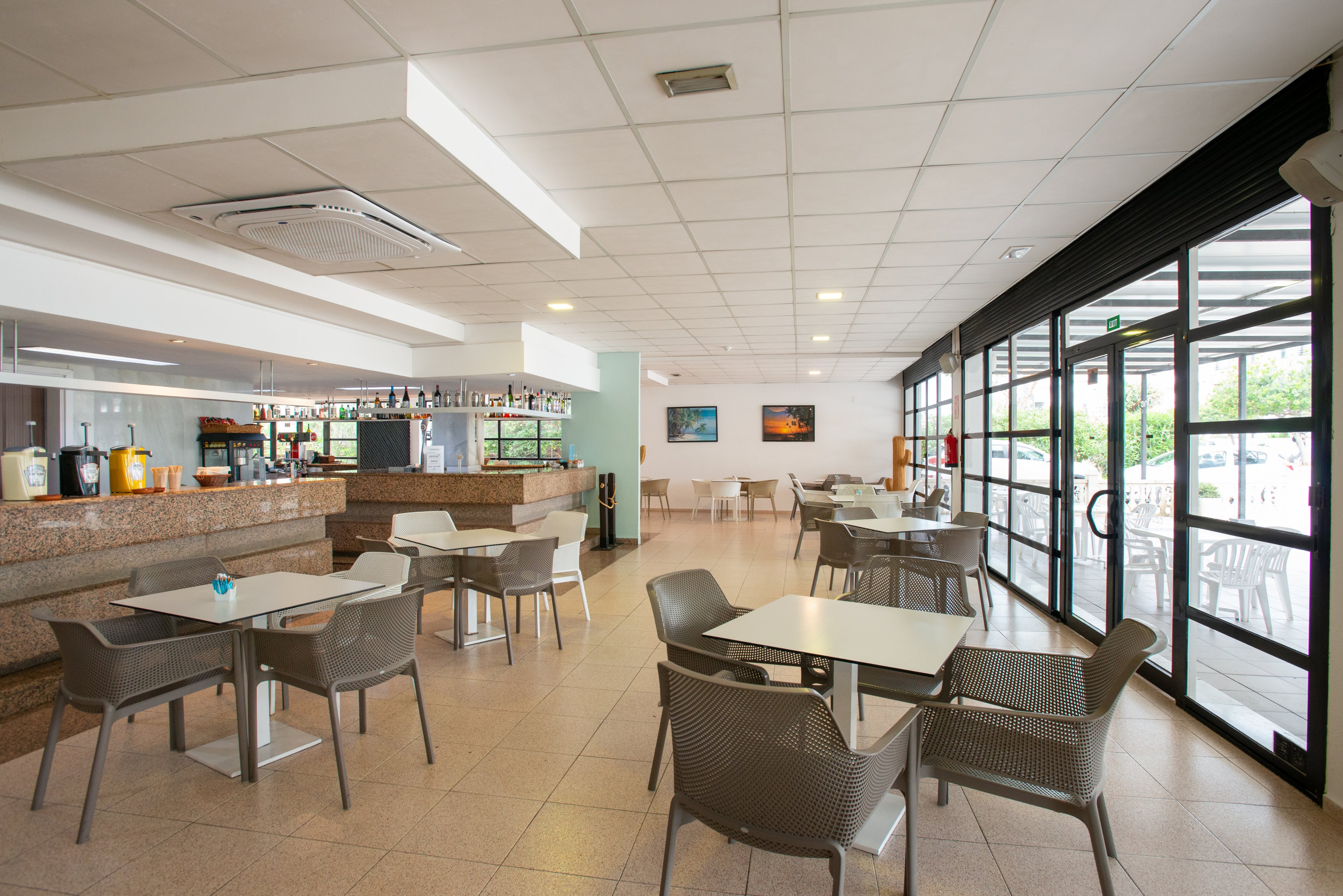 Spacious cafe interior featuring several empty tables and chairs arranged neatly on a tiled floor. A long counter with bar area and beverage dispensers runs along the left side of the room. Large floor-to-ceiling windows and glass doors on the right let in abundant natural light and offer views of greenery outside. The clean, minimalist design and neutral colors create a calm, welcoming atmosphere suitable for dining or casual meetings.
