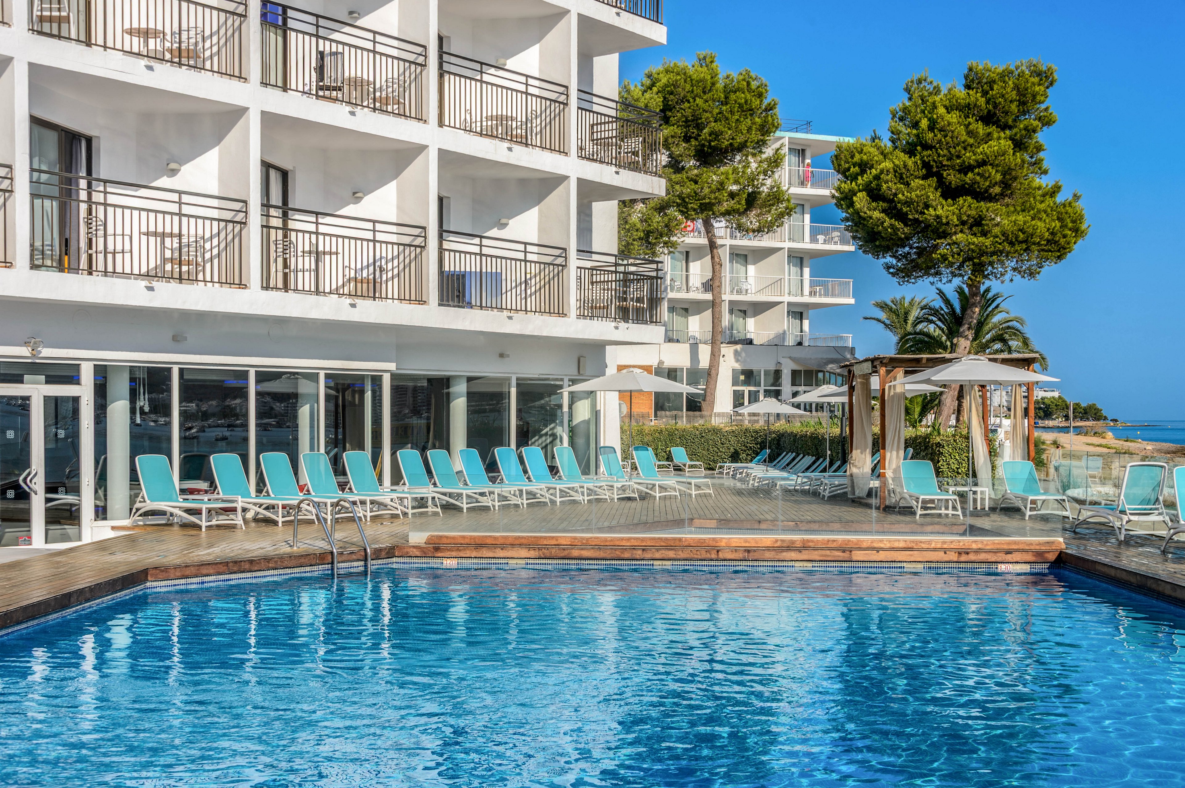 A contemporary multi-story hotel stands beside a clear blue outdoor swimming pool. Rows of turquoise lounge chairs line the pool deck, facing the water and the sea beyond. Large balconies and glass windows overlook the pool area, suggesting guest rooms with ocean views. Tall green trees and a bright blue sky create a relaxed coastal resort atmosphere.