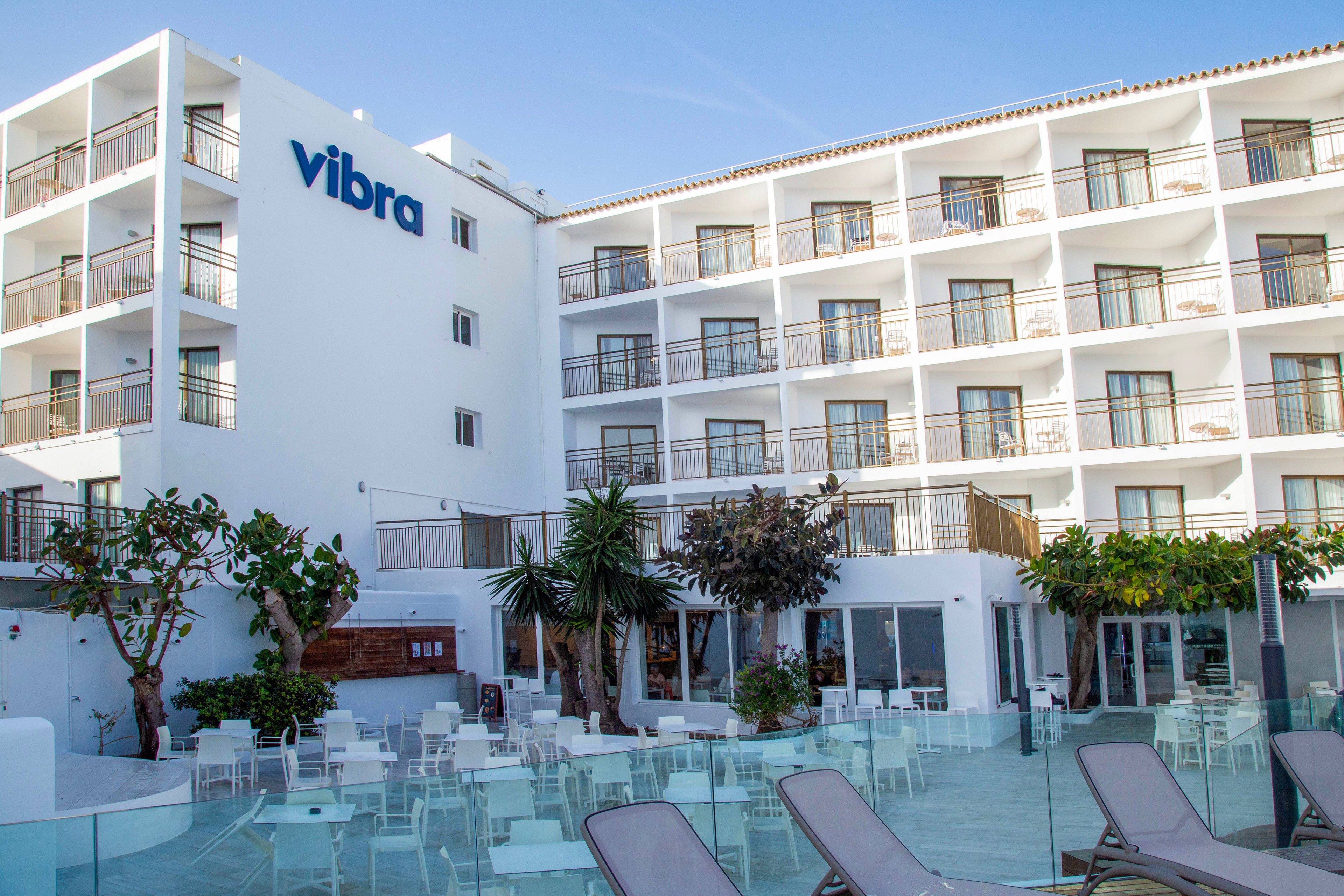 A contemporary white hotel building features multiple floors with balconies and a clean, minimalist facade. The scene overlooks an outdoor pool terrace with glass railings, sun loungers, and neatly arranged tables and chairs. Palm trees and greenery add a touch of nature to the bright, open courtyard. The overall atmosphere suggests a sunny, relaxed vacation setting at a resort property.