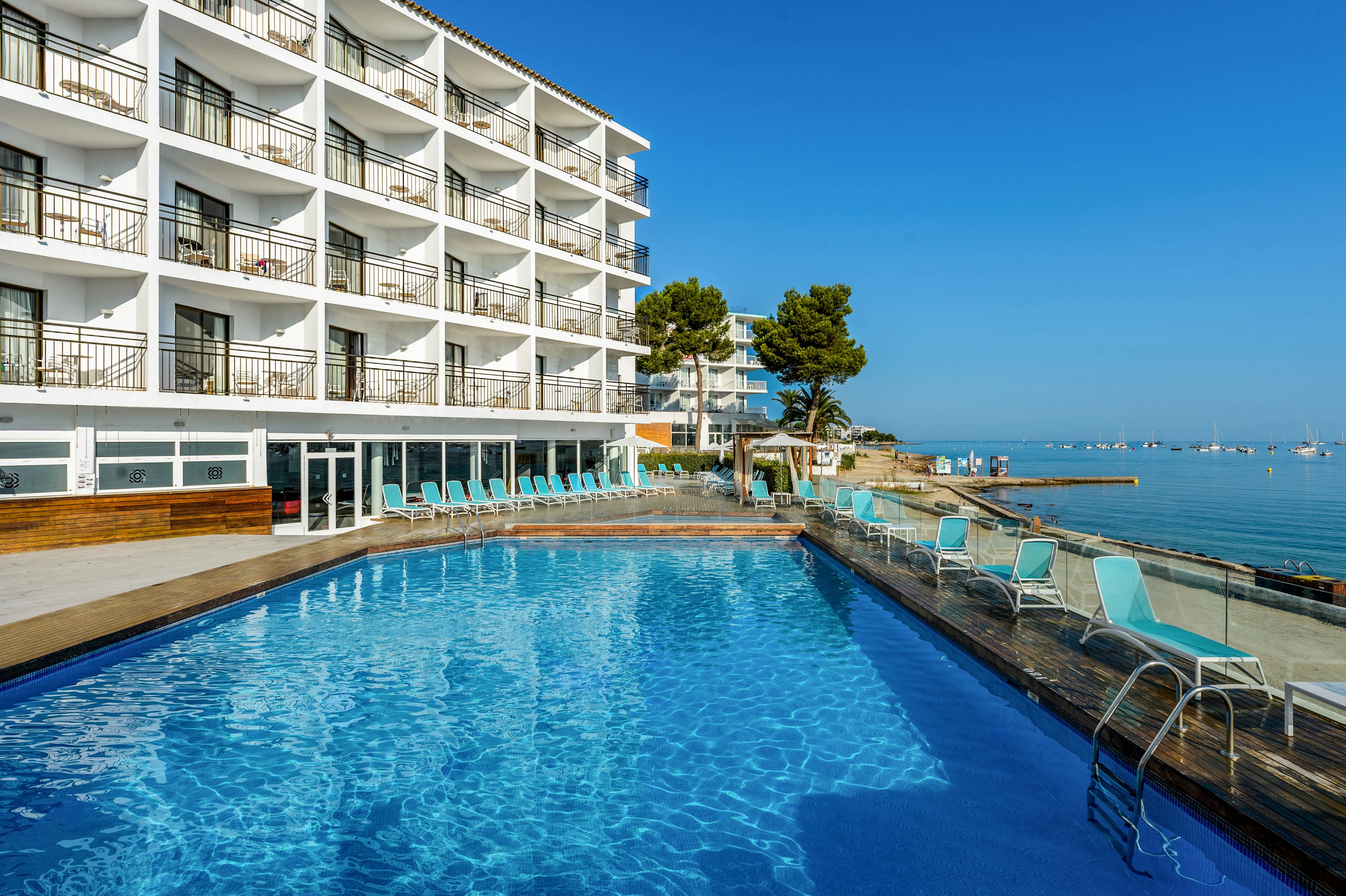 A multi-story seaside hotel overlooks a clear blue outdoor swimming pool. Lounge chairs line the pool deck, facing the calm water and distant boats on the horizon. The building features rows of balconies with railings, offering views of the ocean. Bright sunlight and a cloudless sky create a serene, summery atmosphere ideal for relaxation.