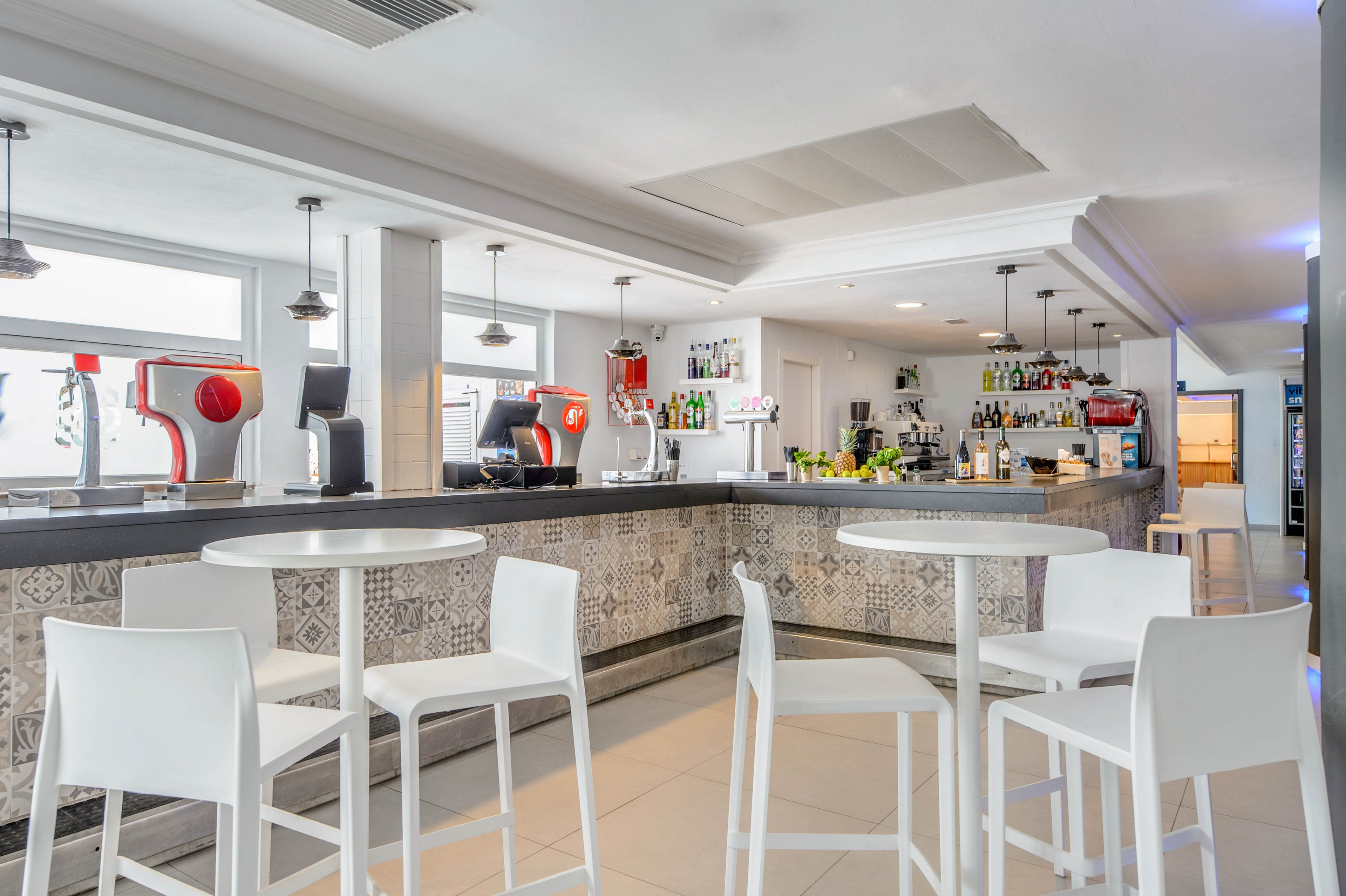 A bright, modern cafe bar interior features high round tables with white stools arranged neatly in the foreground. The long bar counter is decorated with patterned tiles and topped with coffee machines, drink dispensers, and neatly arranged bottles. Pendant lights hang from the ceiling, illuminating the clean, minimalist space. The overall atmosphere is calm and inviting, suggesting a contemporary hospitality setting ready for customers.