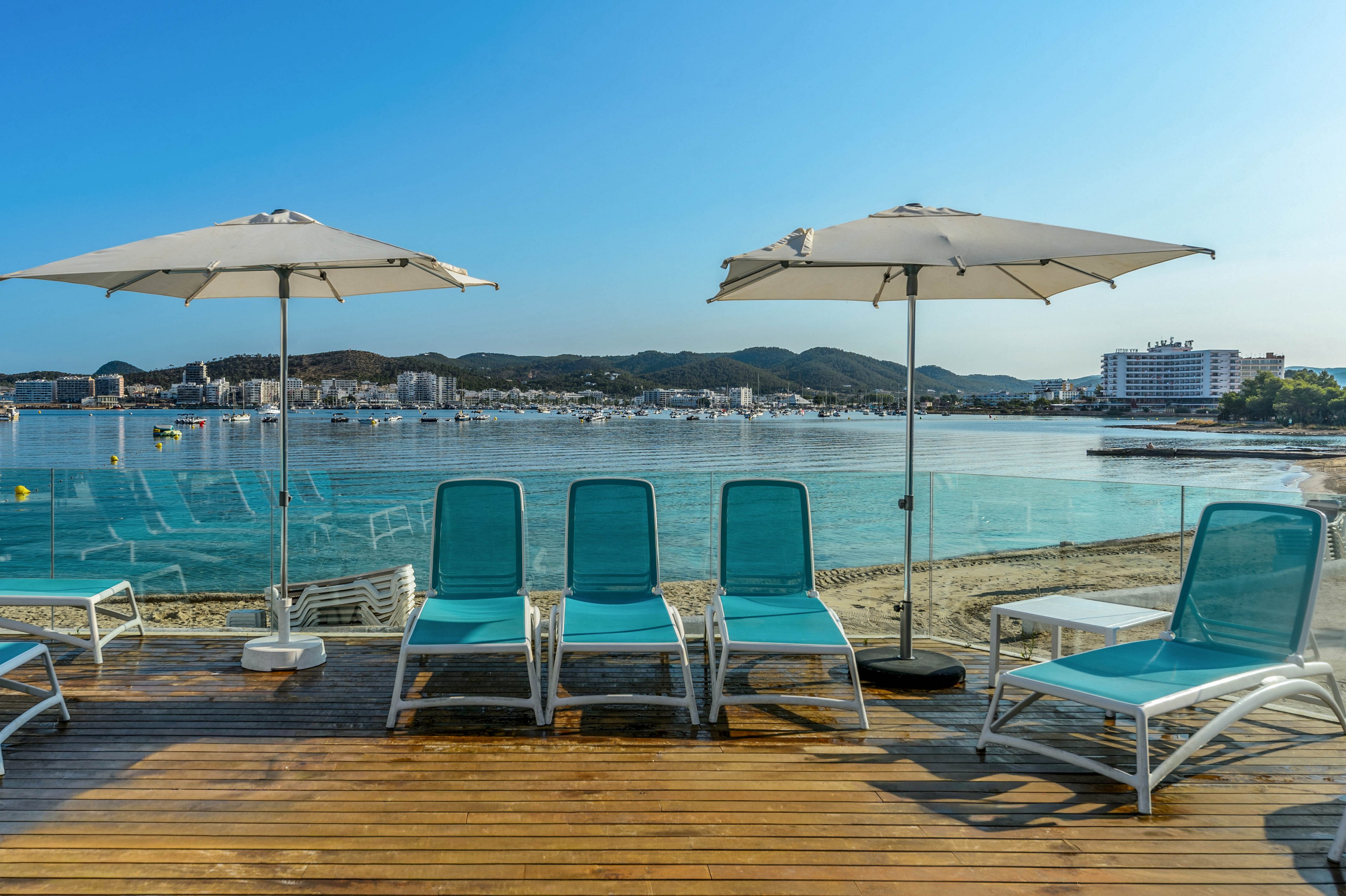 A row of turquoise sun loungers sits on a wooden deck overlooking a calm seaside bay. Two large white umbrellas provide shade over the chairs, while clear blue water stretches out to the distant shoreline. Buildings and boats are visible across the bay under a bright blue sky. The scene suggests a relaxing vacation setting at a coastal resort.