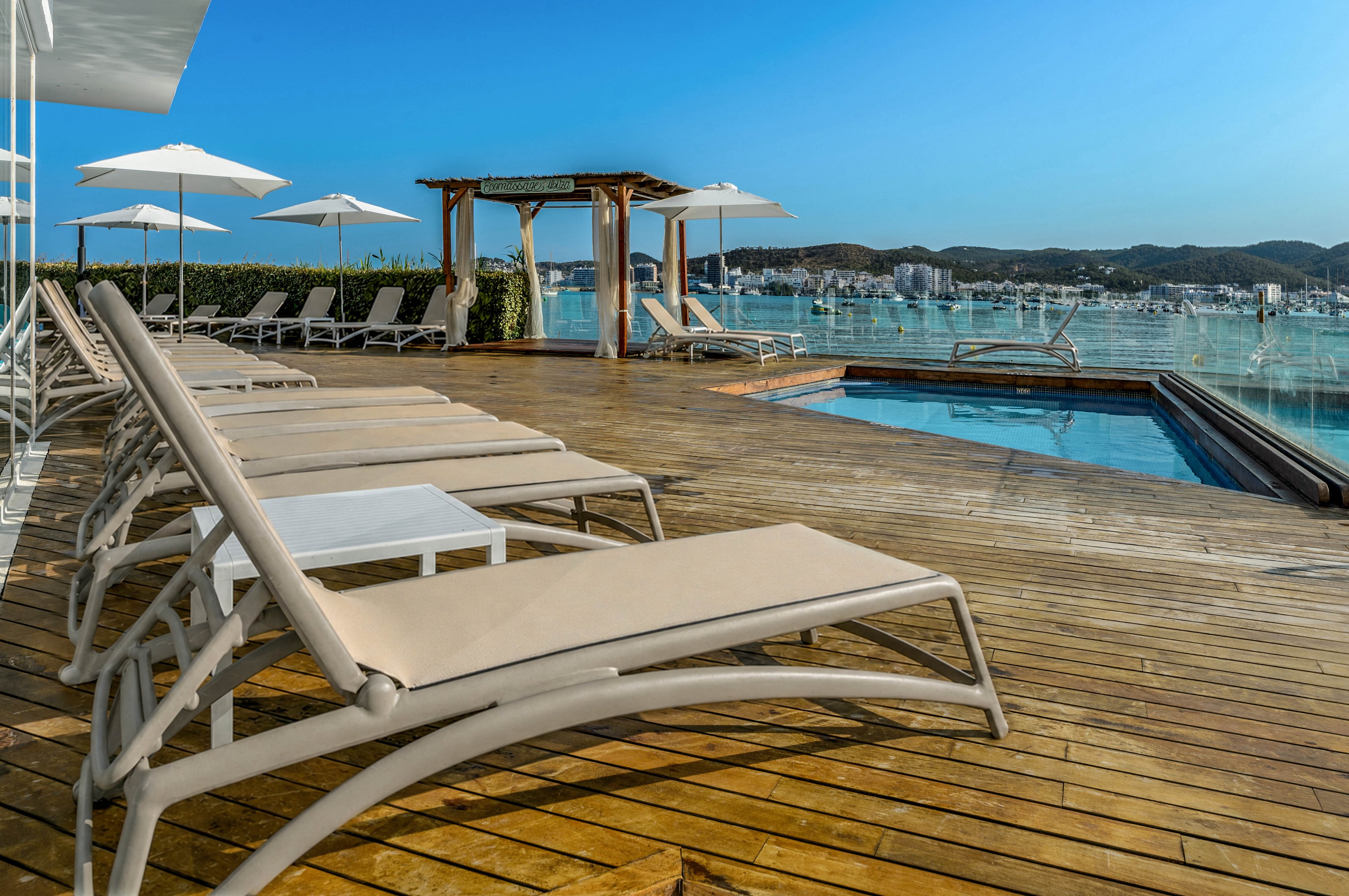 A contemporary rooftop pool area features several beige sun loungers arranged on a wooden deck. White umbrellas provide shade along the edge, while a small pergola structure frames the view toward the horizon. The clear blue swimming pool reflects the bright sky and surrounding glass railings. In the distance, a coastal cityscape and hills create a relaxed vacation atmosphere.