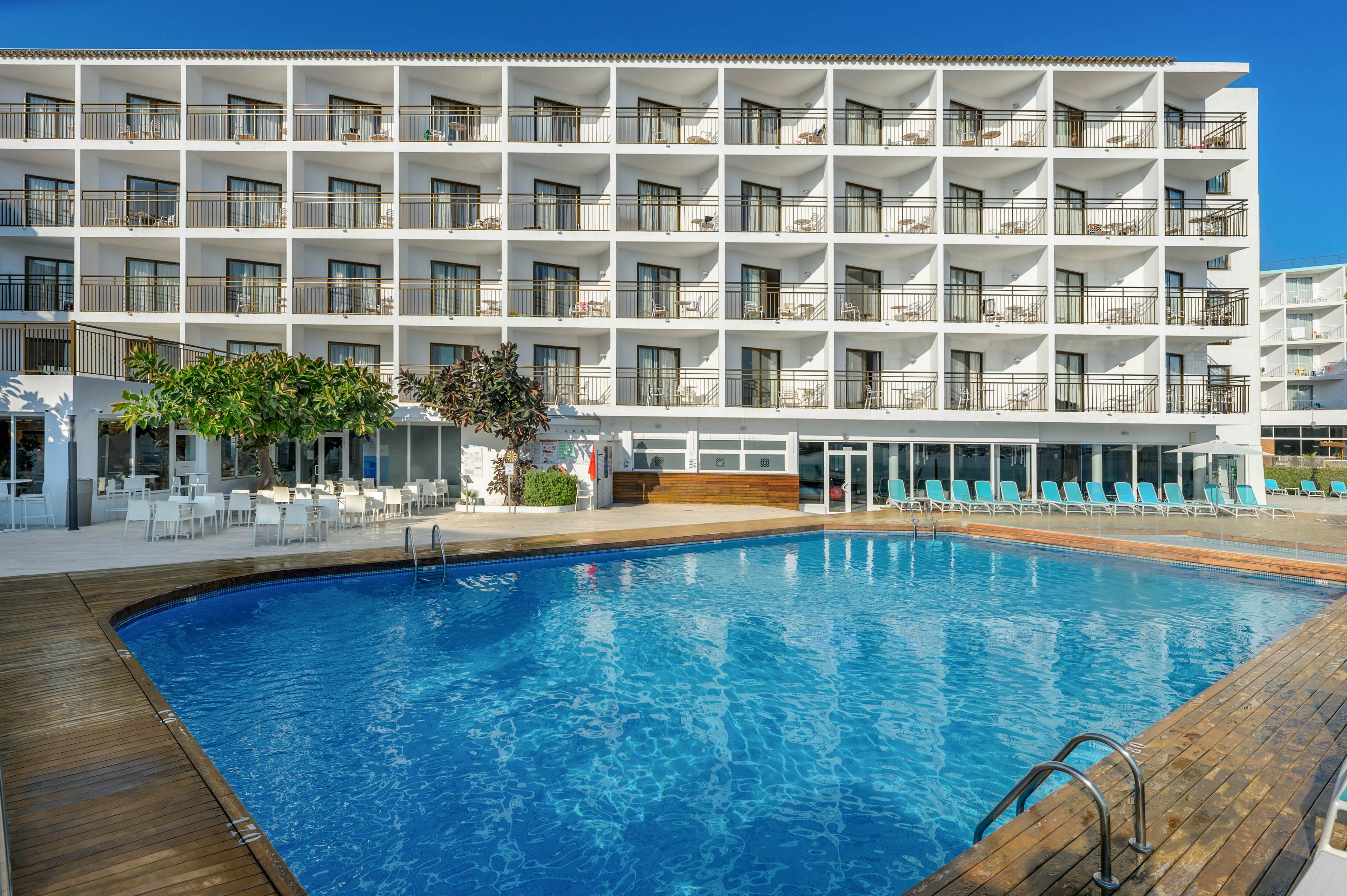 A multi-story hotel building with rows of balconies overlooks a large outdoor swimming pool. The pool is surrounded by wooden decking and metal ladders leading into the clear blue water. Sun loungers are lined up near the glass-fronted ground floor, suggesting a relaxing resort atmosphere. Bright daylight and clear skies enhance the clean, modern appearance of the property.