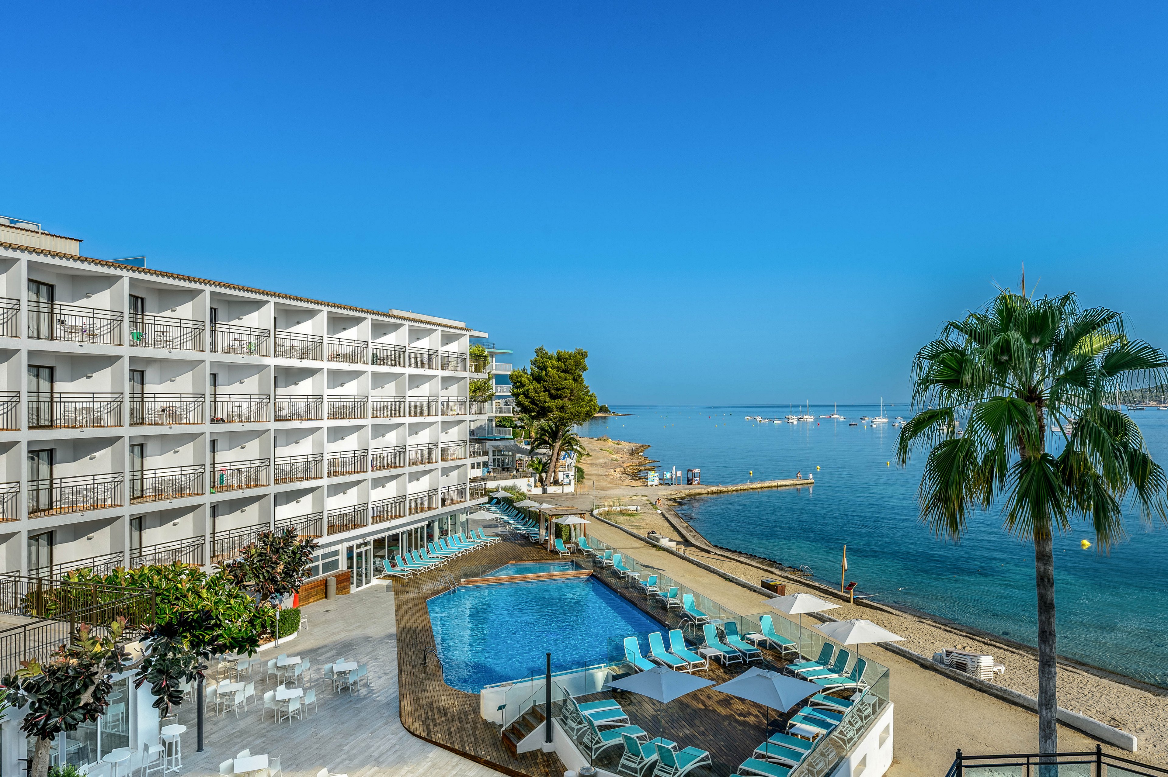 A modern multi-story seaside hotel overlooks a bright blue swimming pool and a calm shoreline. Sun loungers and umbrellas surround the pool area, creating a relaxed resort atmosphere. The scene opens onto a sandy beach and a clear, tranquil sea dotted with small boats and a distant marina. A tall palm tree and lush greenery add a tropical touch under a clear blue sky.