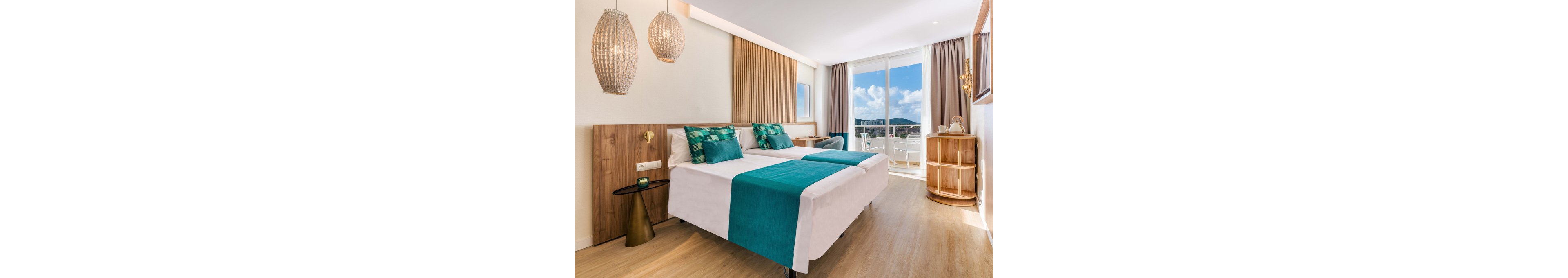A contemporary hotel room features two neatly made twin beds with white linens and teal accents. Wooden wall panels, woven pendant lights, and minimalist furniture create a warm, modern aesthetic. Large glass doors open onto a balcony with outdoor seating and a scenic view of the coastline and blue sky. Natural light fills the space, highlighting the clean, inviting atmosphere.