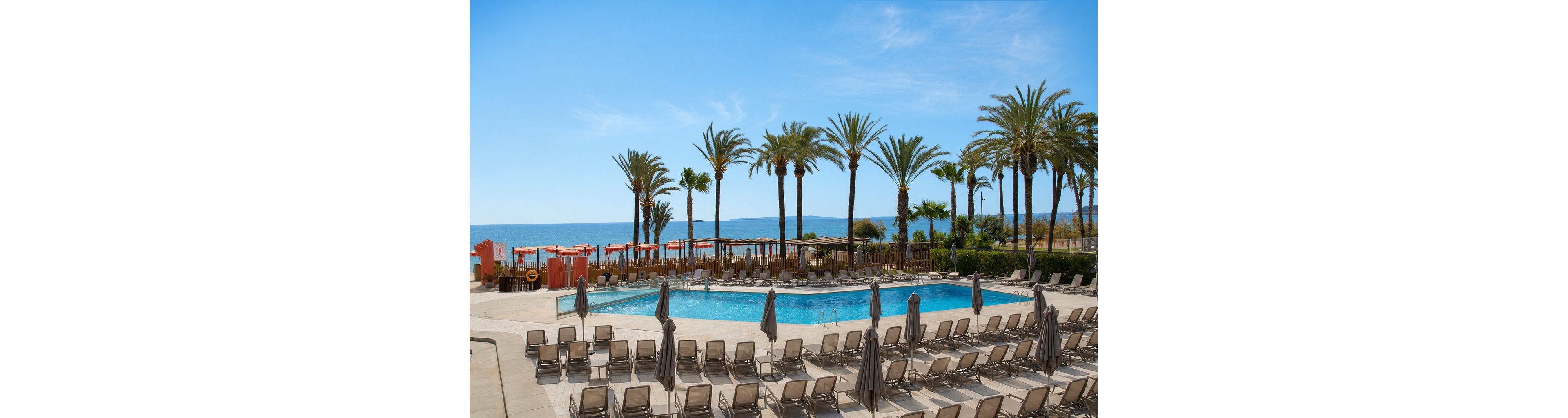 An outdoor swimming pool sits at the center of a beachfront resort lined with tall palm trees. Numerous lounge chairs and closed umbrellas surround the pool, facing toward the ocean. Orange cabanas and parasols add color along the water’s edge, while the calm sea stretches out under a clear blue sky. The scene suggests a warm, relaxing vacation destination with ample seating and open space.