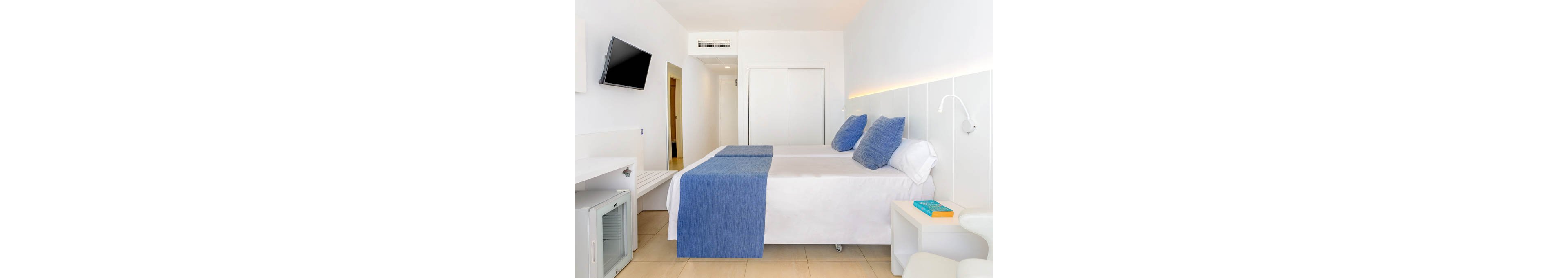 A modern hotel room features a neatly made double bed with blue pillows and a matching blue runner. The space is predominantly white, including the walls, furniture, and tiled floor, creating a clean and minimalist look. A flat-screen TV is mounted on the wall opposite the bed, and a small desk area with a mini fridge sits to the side. Soft ambient lighting along the wall and a bedside table with a book add a touch of comfort to the contemporary setting.
