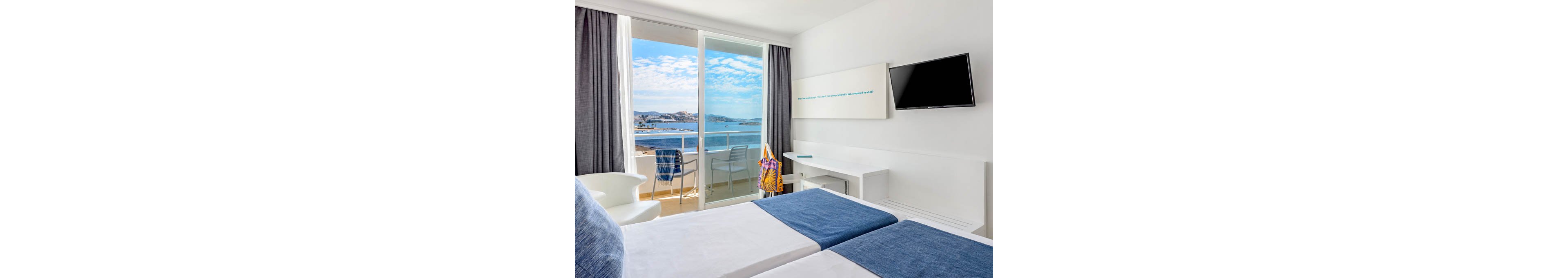Bright modern hotel room with twin beds overlooks a scenic coastal bay. Large glass doors open onto a small balcony with chairs and a table facing the ocean. The interior features white walls, blue bed runners, a wall-mounted television, and a minimalist desk area. A colorful beach bag hangs on the balcony chair, suggesting a relaxed vacation atmosphere under a clear blue sky.