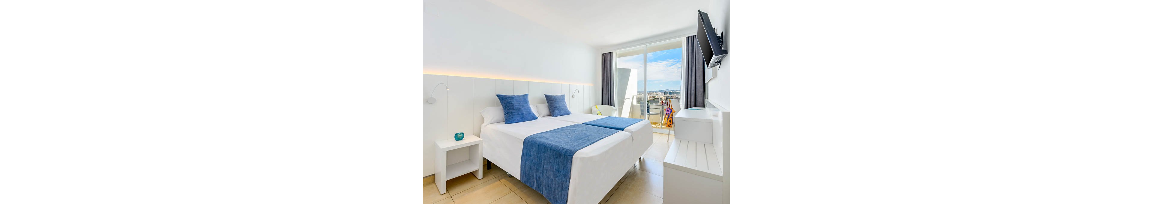 A contemporary hotel room features a double bed with crisp white linens and blue accent pillows and runner. The space is minimalistic, with white walls, light tiled flooring, and simple bedside furniture. Large glass doors open onto a balcony overlooking a coastal cityscape under a blue sky. A wall-mounted television, sheer and dark curtains, and a small decorative teal candle add to the clean, relaxed atmosphere.