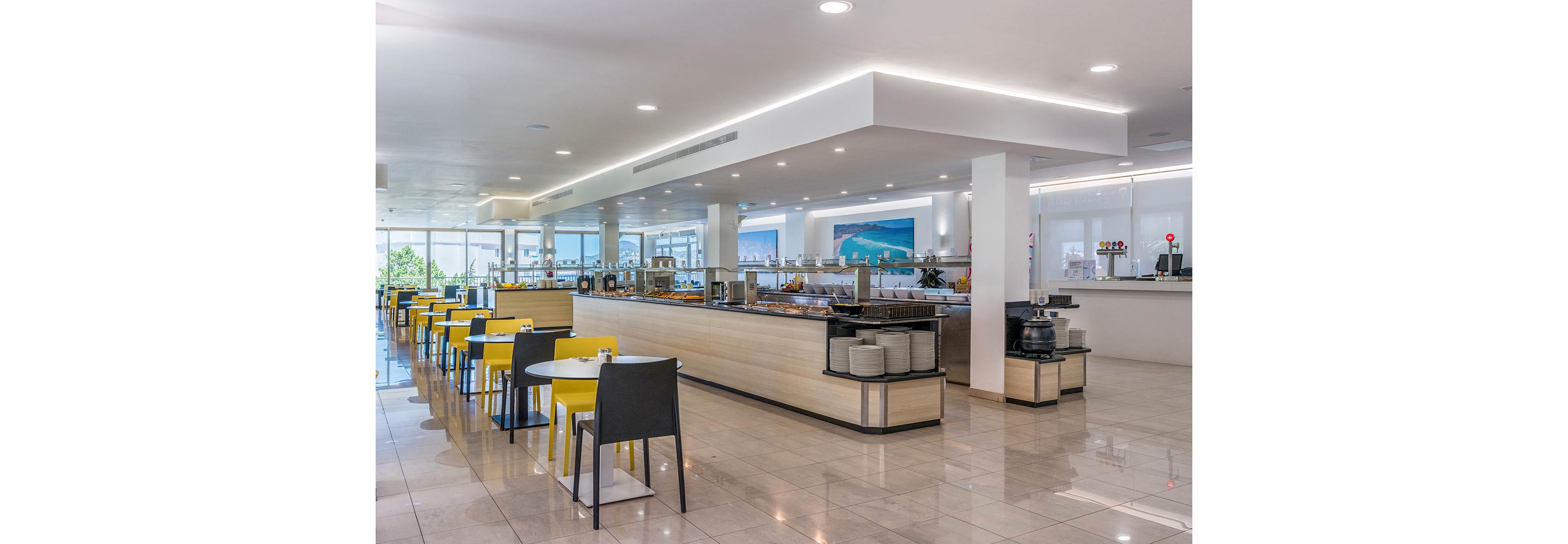 Bright, contemporary self-service restaurant with long buffet counters dominates the scene. Yellow and black chairs surround neatly arranged tables on a polished tiled floor. Large windows at the back let in natural daylight and reveal an outdoor view. The space appears clean, spacious, and ready for guests, with dishes and beverage stations prepared along the counters.