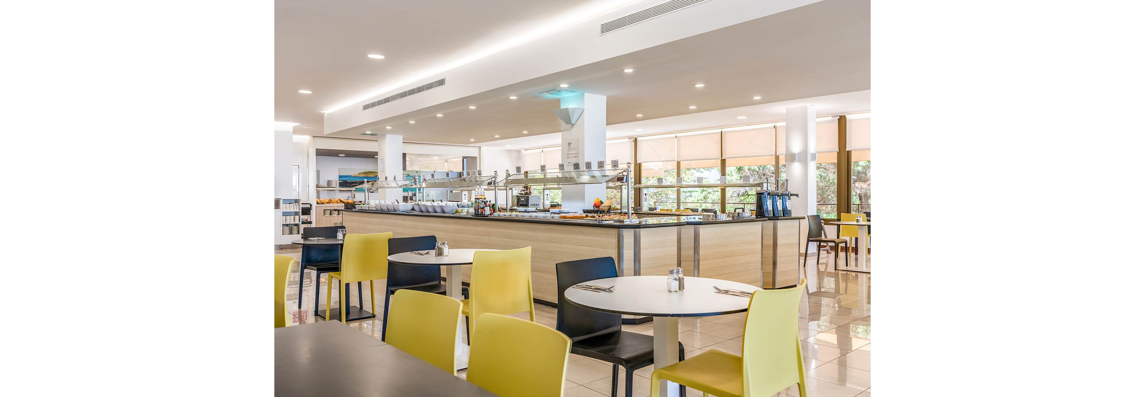 Bright, contemporary restaurant interior featuring a central self-service buffet counter. Empty round tables with yellow, white, and black chairs are arranged on a glossy tiled floor. Large windows with beige blinds let in natural light and offer views of greenery outside. Recessed ceiling lights and clean white walls create a spacious, minimalist dining atmosphere.