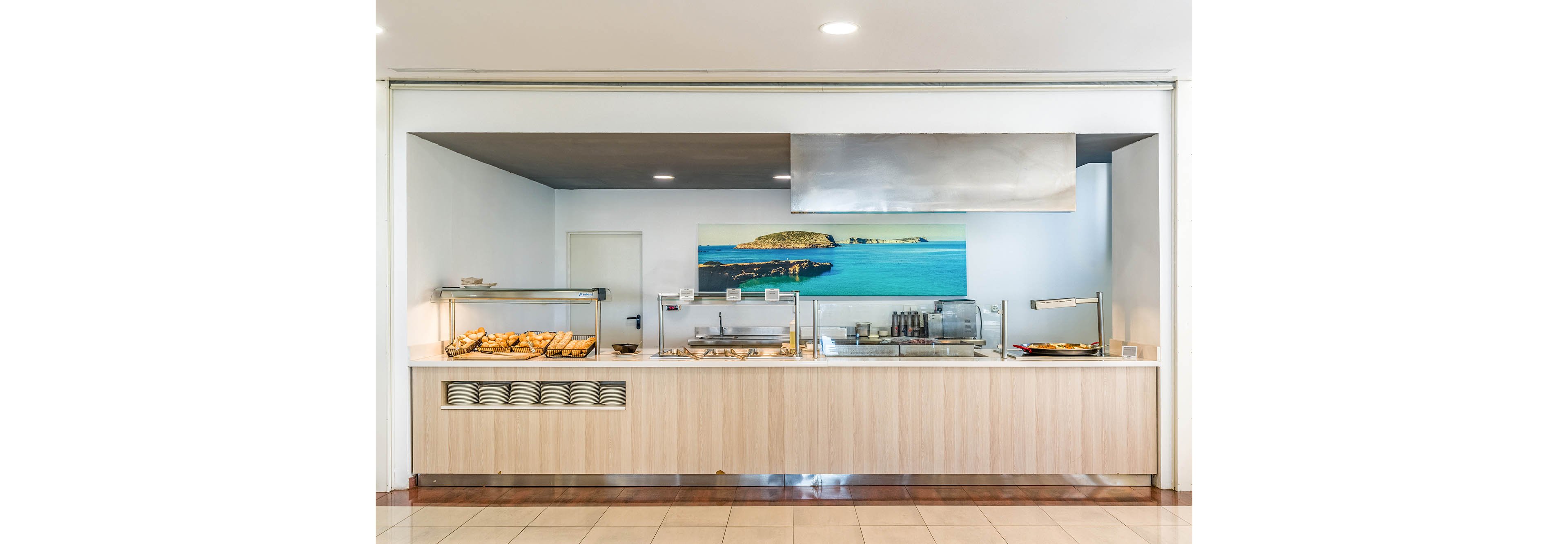 A clean, modern self-service buffet counter is set up in a cafeteria or hotel dining area. Trays of bread, pastries, and hot dishes are displayed under warming lamps along the light wood counter. Stacks of white plates are neatly arranged on the lower shelf, ready for guests. A large seascape photograph decorates the back wall, adding a bright blue accent to the otherwise neutral interior.