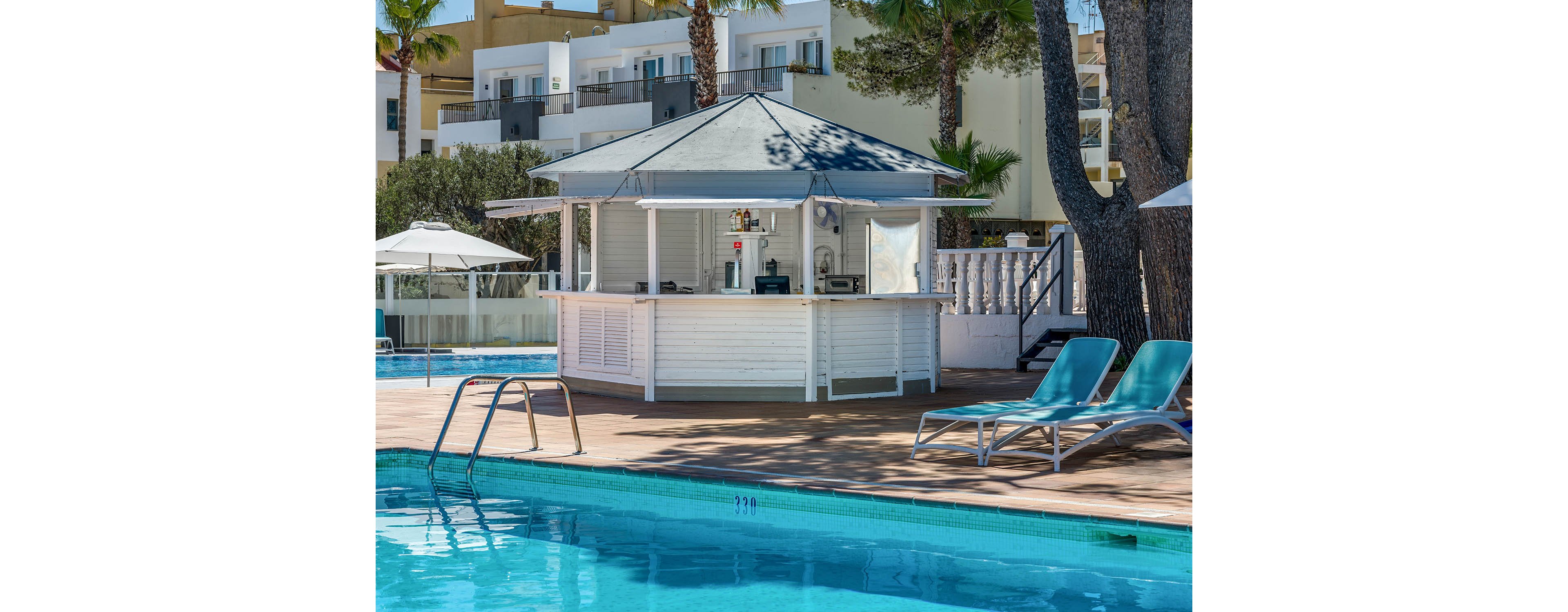 An outdoor poolside bar stands beside a clear blue swimming pool at a modern resort. The white kiosk-style bar is shaded by a roof and stocked with drinks and equipment. Two turquoise sun loungers sit on the deck near the water, with trees and multi-story buildings in the background. Bright daylight and calm water create a relaxed vacation atmosphere. The pool edge shows depth markings along the tiled border.