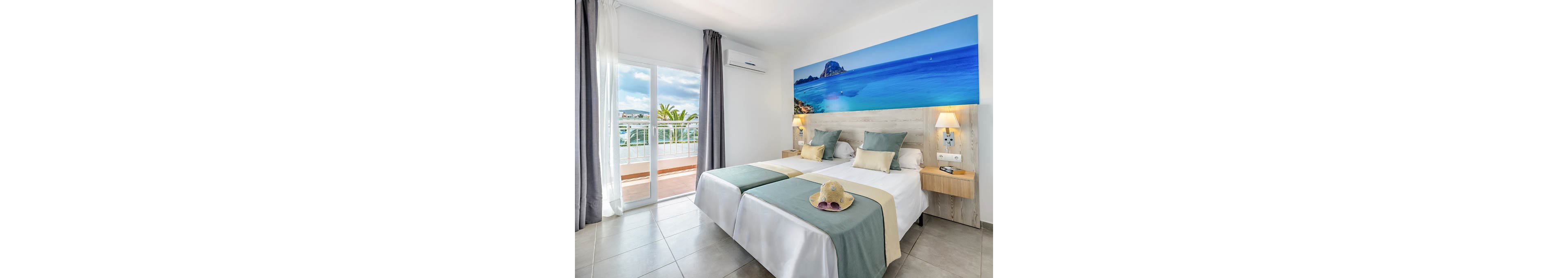 Modern twin beds are arranged neatly in a bright hotel room with soft green runners and beige accent pillows. A large panoramic seascape artwork above the headboard highlights a vivid blue ocean and rocky coastline. Sunlight streams through the glass doors leading to a balcony with white railings and a view of palm trees and distant buildings. Neutral tiled flooring, bedside lamps, and a wall-mounted air conditioner complete the clean, contemporary interior.