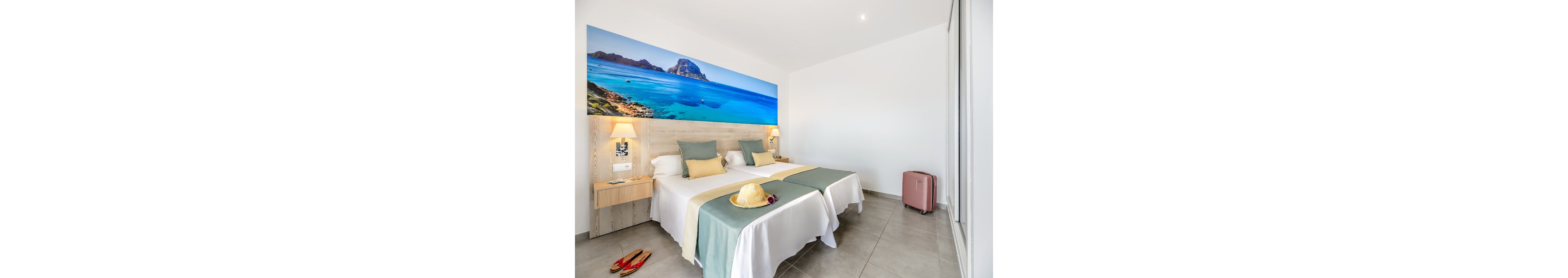 A contemporary hotel room features two neatly made twin beds with white linens and soft pastel throws. Above the headboard, a large panoramic photograph of a tropical coastline adds vibrant color to the space. Nightstands with wall-mounted lamps flank the beds, while a pink suitcase stands near the wardrobe and a straw hat and sandals suggest a vacation stay. The room is clean, minimal, and filled with natural light, creating a relaxed holiday atmosphere.