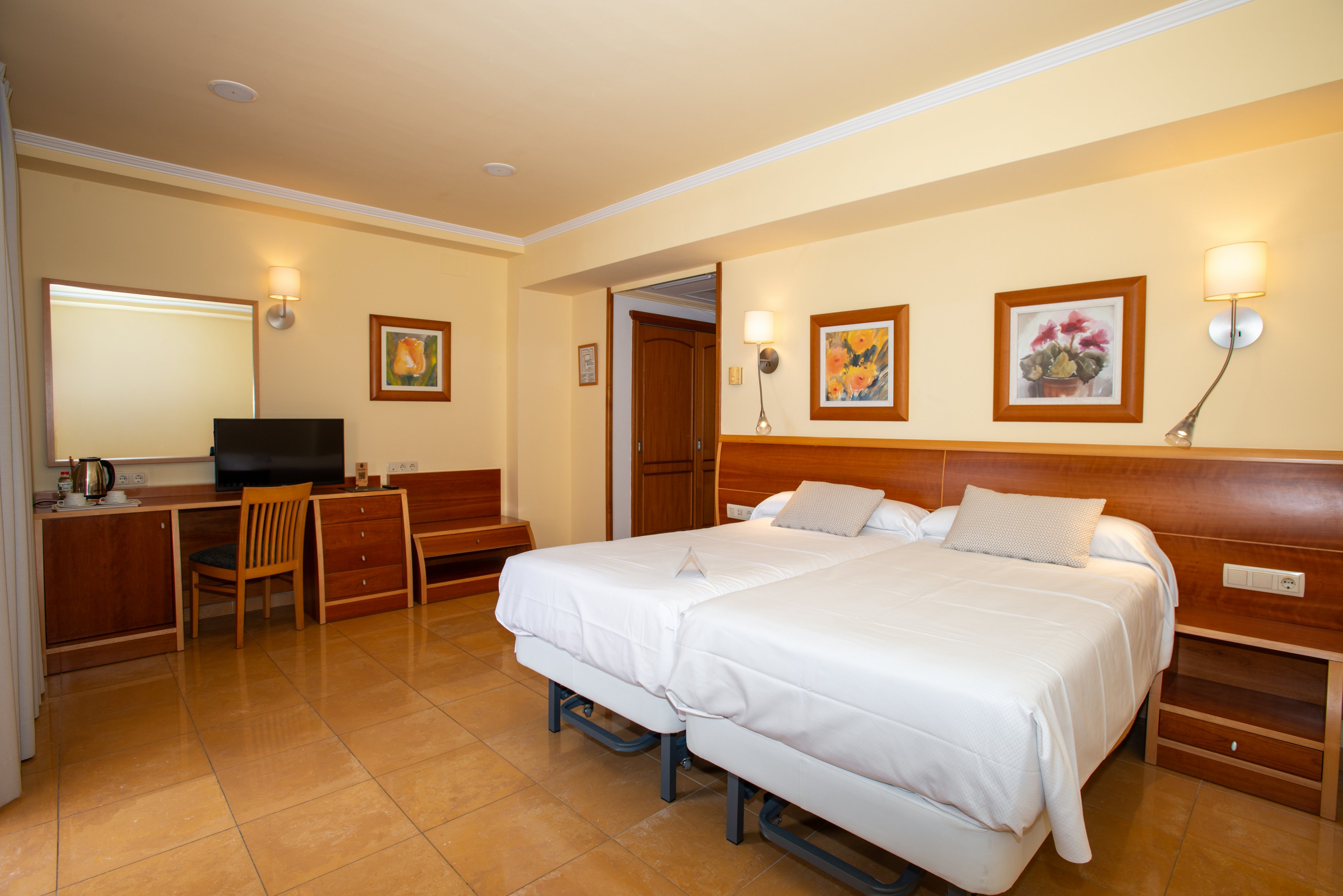A tidy hotel room features two neatly made twin beds pushed together with white linens and neutral pillows. The space includes a wooden desk with a chair, a flat-screen television, and a small beverage station with a kettle. Warm yellow walls are decorated with framed artwork and modern wall-mounted lamps that create a cozy ambiance. The room has a clean tiled floor and simple, functional furniture suitable for travelers.