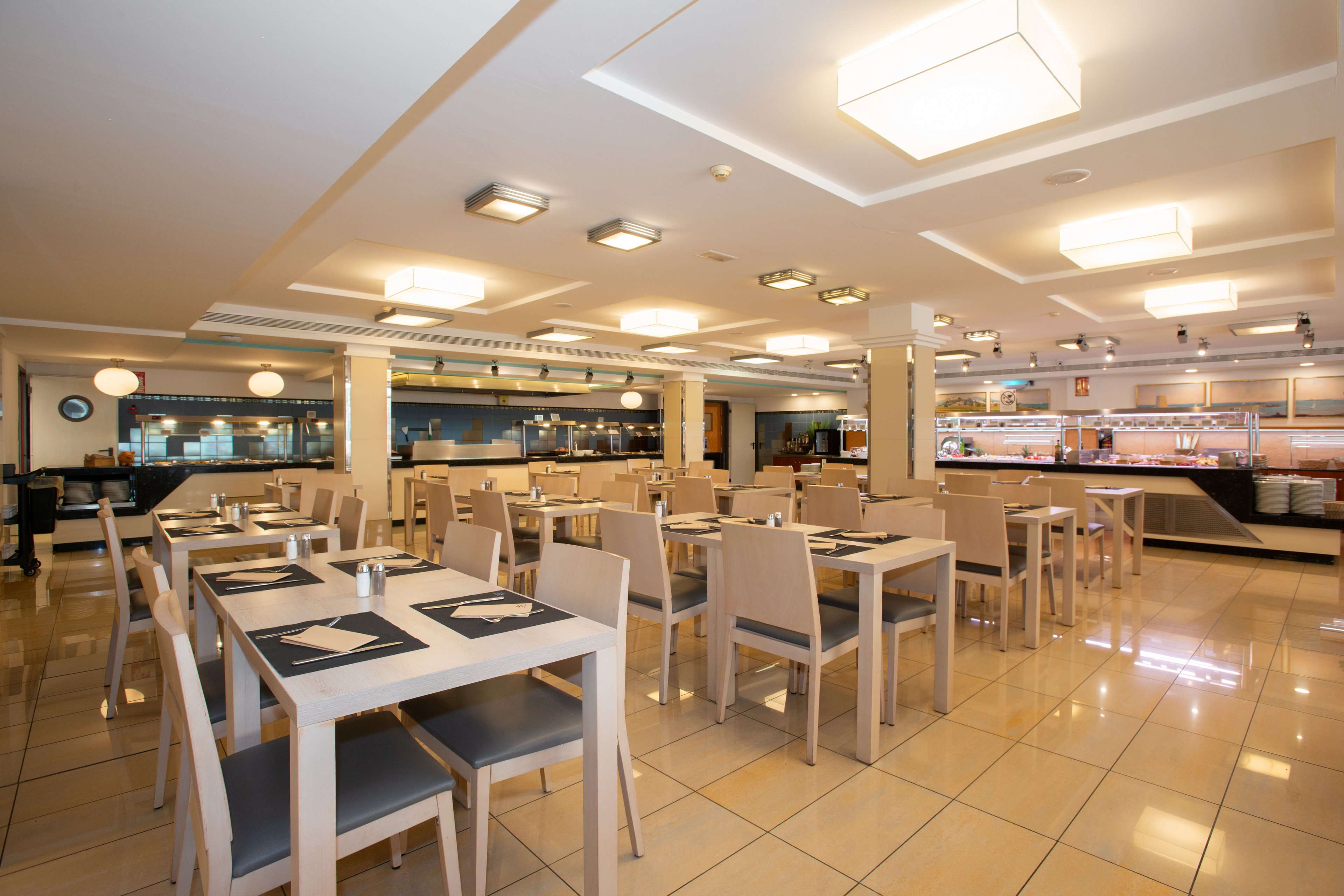 Spacious modern restaurant interior with neatly arranged tables and chairs. The dining area features light-colored furniture, tiled floors, and large ceiling lights that create a bright atmosphere. Place settings with dark placemats and cutlery are prepared on each table. In the background, an open kitchen and buffet counter are visible, suggesting a self-service or hotel restaurant setting.