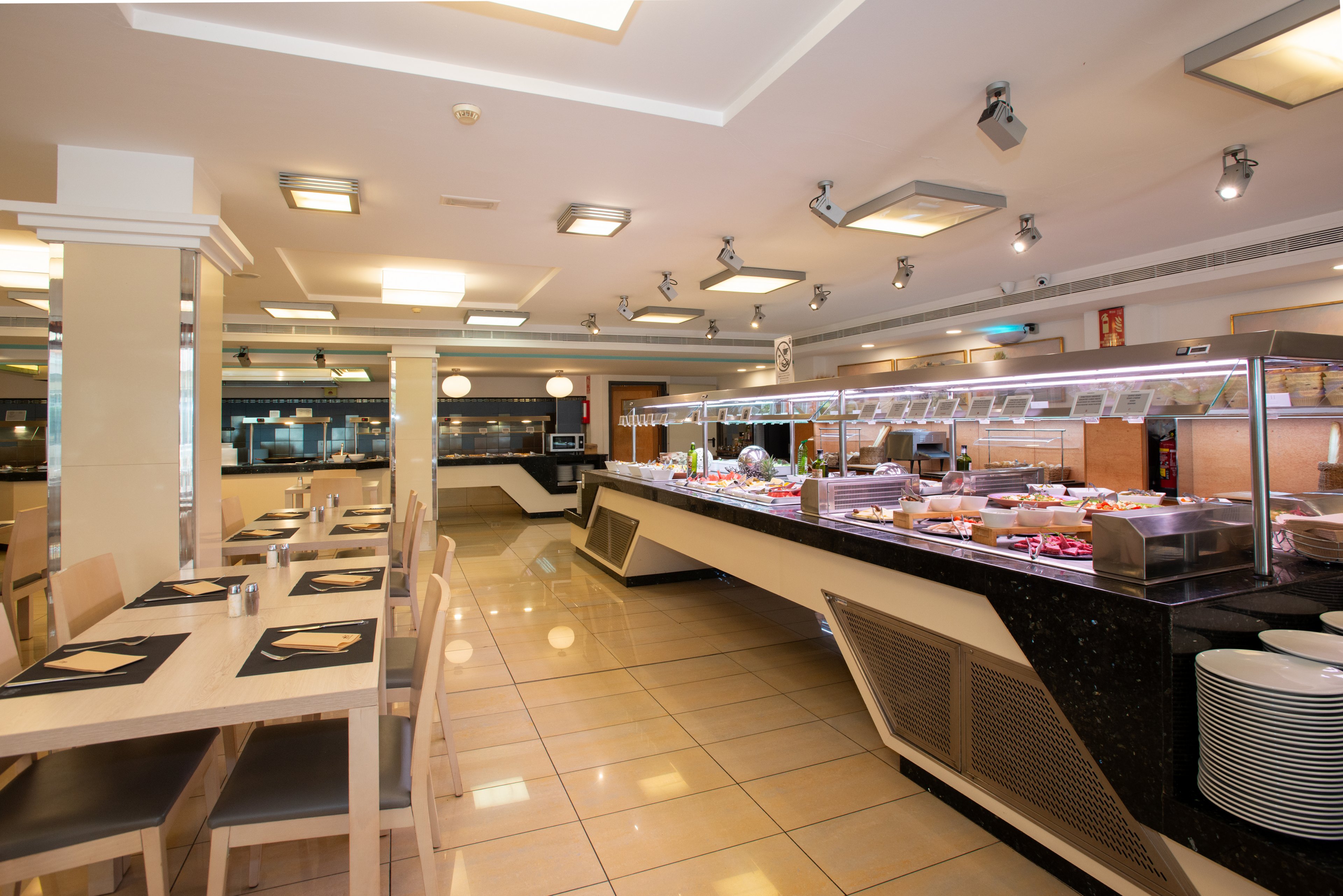 Spacious self-service buffet area in a modern restaurant dining room. A long counter displays various foods in stainless steel trays under a glass sneeze guard. Wooden tables and chairs with black placemats line the tiled floor, creating an orderly seating area. Bright ceiling lights and neutral colors give the space a clean, contemporary atmosphere. The overall setting suggests a hotel or cafeteria-style dining experience.