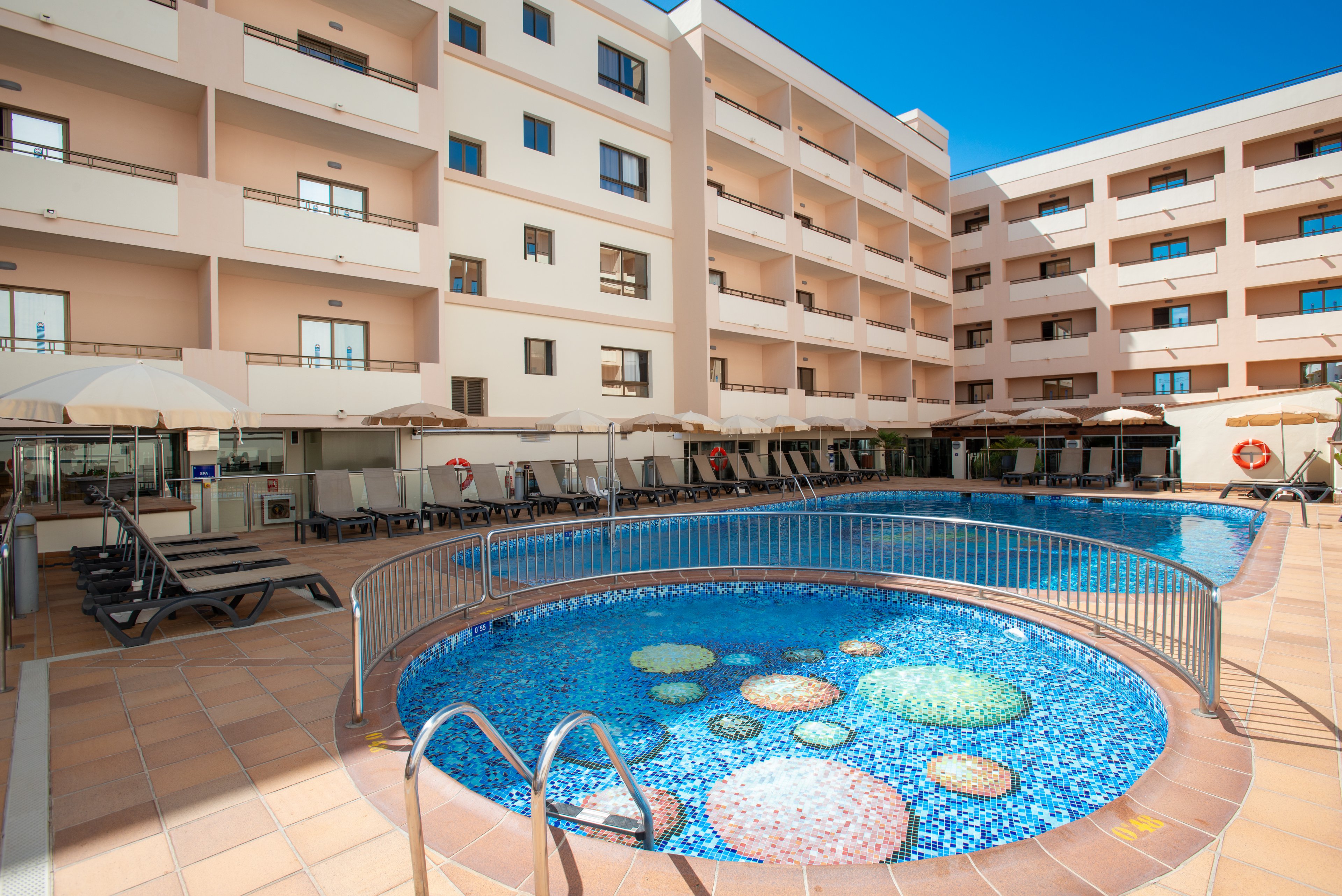 A modern hotel courtyard features a circular shallow pool in the foreground and a larger curved swimming pool behind it. The smaller pool has a colorful mosaic floor with round patterns and is surrounded by a metal safety railing. Sun loungers and umbrellas line the tiled pool deck, suggesting a relaxing vacation setting. Multi-story beige apartment-style buildings frame the scene under a clear blue sky.