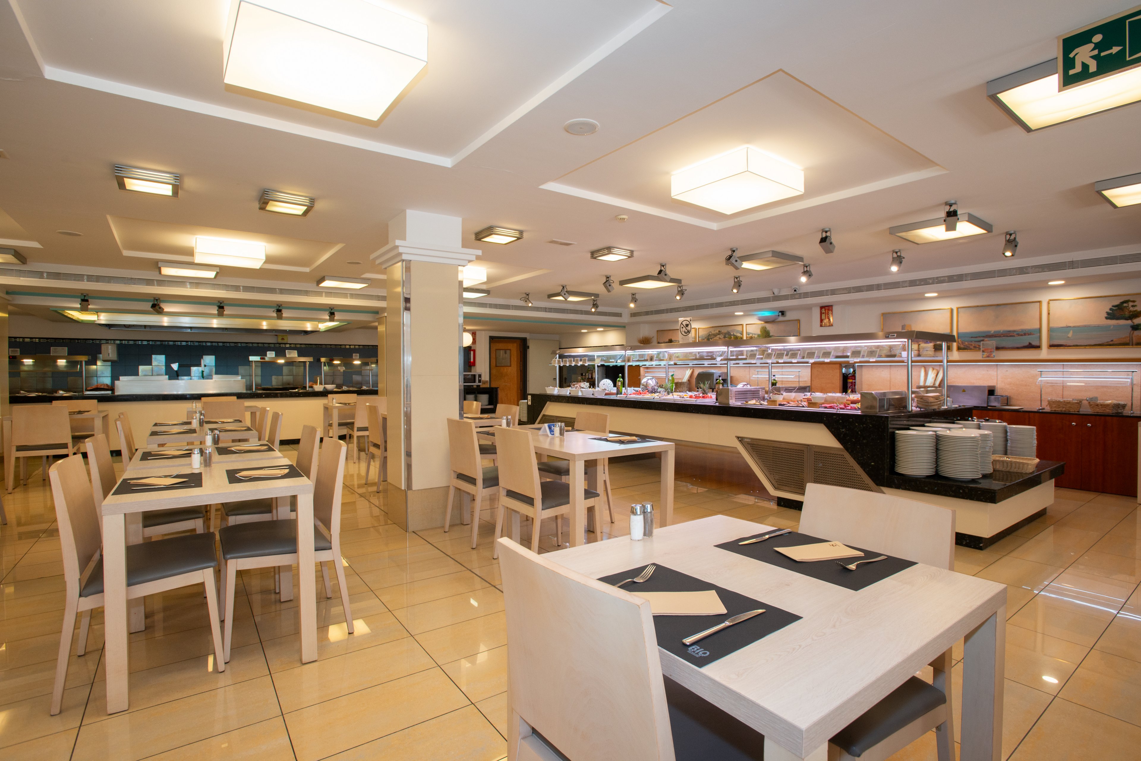 Spacious modern restaurant interior featuring light wooden tables and chairs arranged neatly on a tiled floor. The setting includes a long self-service buffet counter with various food items displayed under bright lighting. Ceiling-mounted square lights and clean lines give the space a contemporary, minimalist feel. The restaurant appears empty, emphasizing the orderly layout and inviting atmosphere.