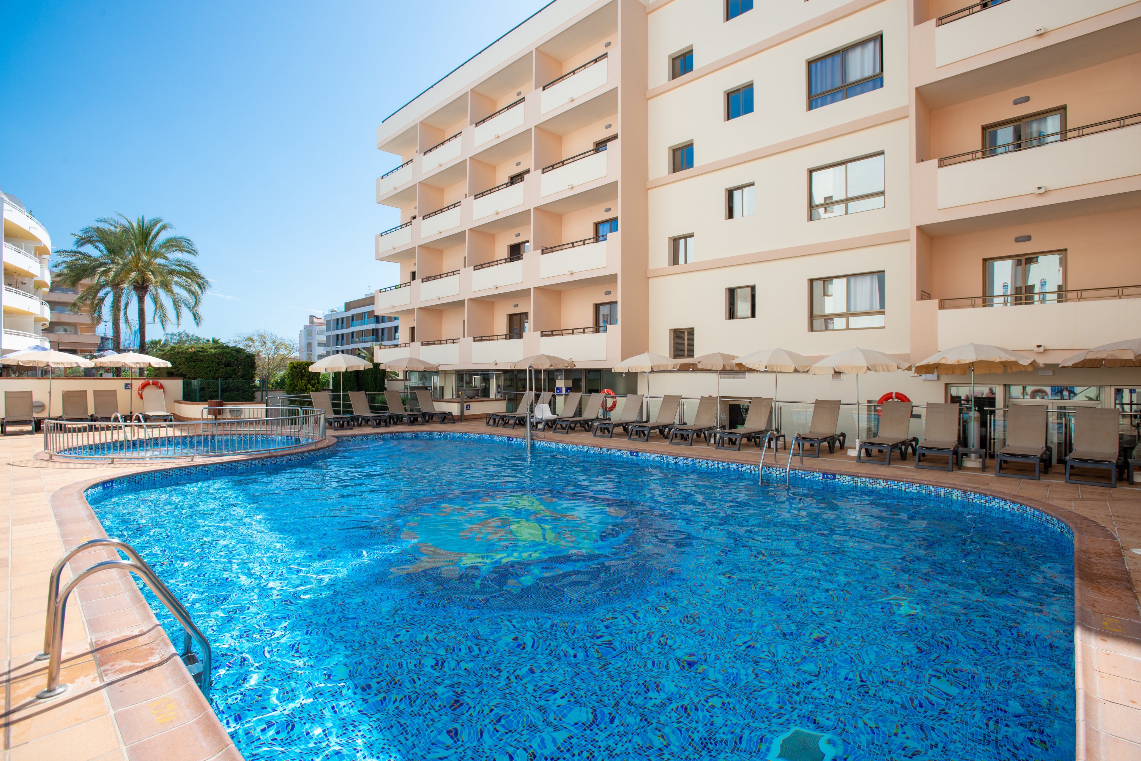 A large outdoor swimming pool sits in the courtyard of a multi-story hotel building. Sun loungers and umbrellas line the tiled pool deck, creating a relaxing resort atmosphere. The clear blue water reflects the bright sky, and a palm tree is visible near the edge of the property. Balconies overlook the pool area, suggesting guest rooms with pool views.