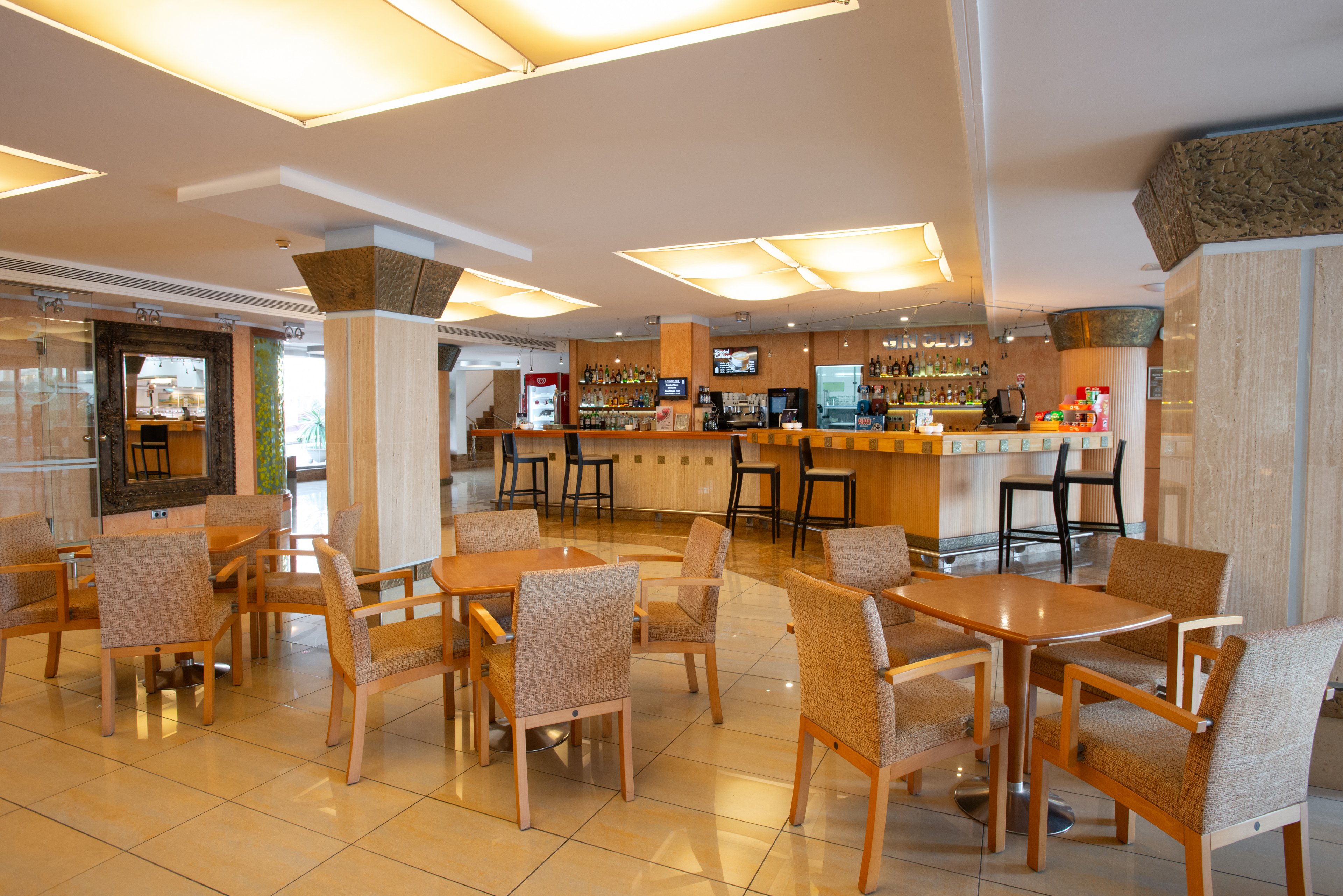 Spacious hotel lobby bar featuring several wooden tables with woven chairs neatly arranged on a polished tile floor. The bar counter in the background is stocked with bottles and glassware, with high stools lined along the front. Warm ceiling lights and neutral-toned columns create a welcoming, contemporary atmosphere. Large windows and glass doors suggest access to an outdoor area, enhancing the open, airy feel of the space.