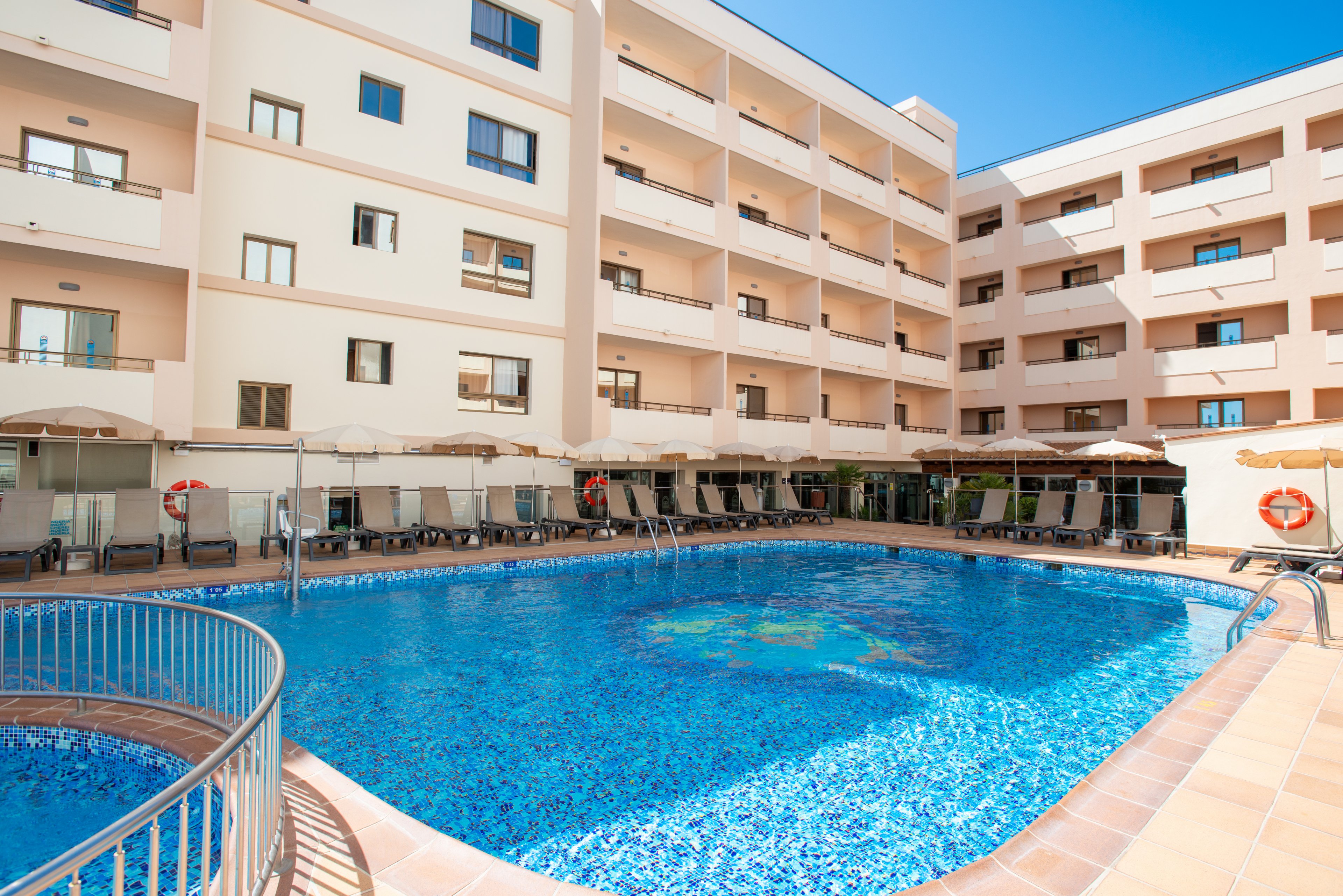 A large outdoor swimming pool sits in the courtyard of a modern multi-story hotel building. Sun loungers and umbrellas line the poolside terrace, suggesting a relaxing vacation setting. The clear blue water reflects the bright sky, creating a vibrant and inviting atmosphere. Balconies overlook the pool area, emphasizing resort-style accommodation and leisure.