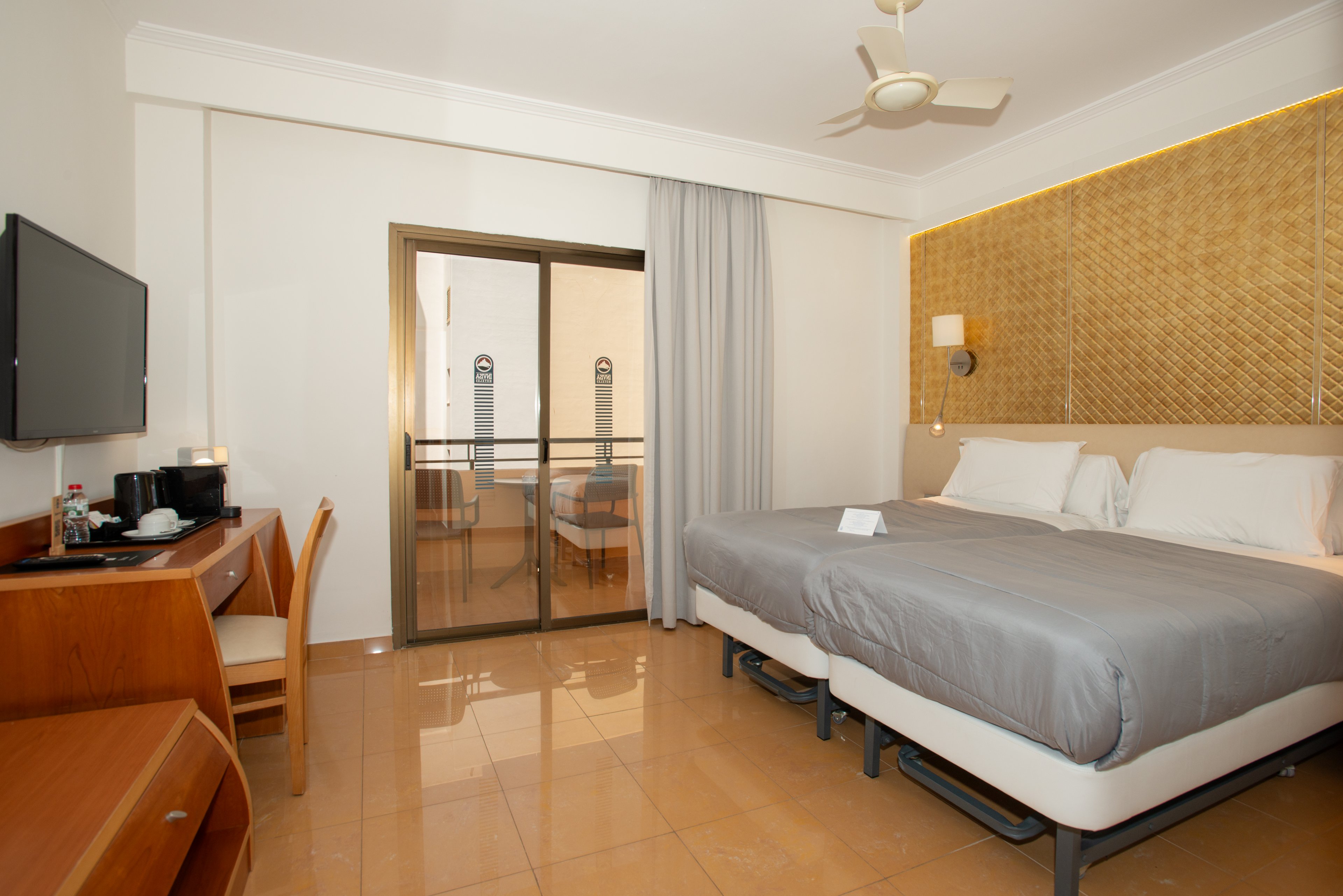 A bright hotel room features two neatly made twin beds with gray bedding against a textured accent wall. A sliding glass door opens to a small balcony with outdoor chairs and a view of neighboring buildings. On the left, a wooden desk holds a flat-screen television, coffee maker, cups, and bottled water. Warm lighting, tiled floors, and neutral colors create a clean, contemporary atmosphere suitable for travelers.