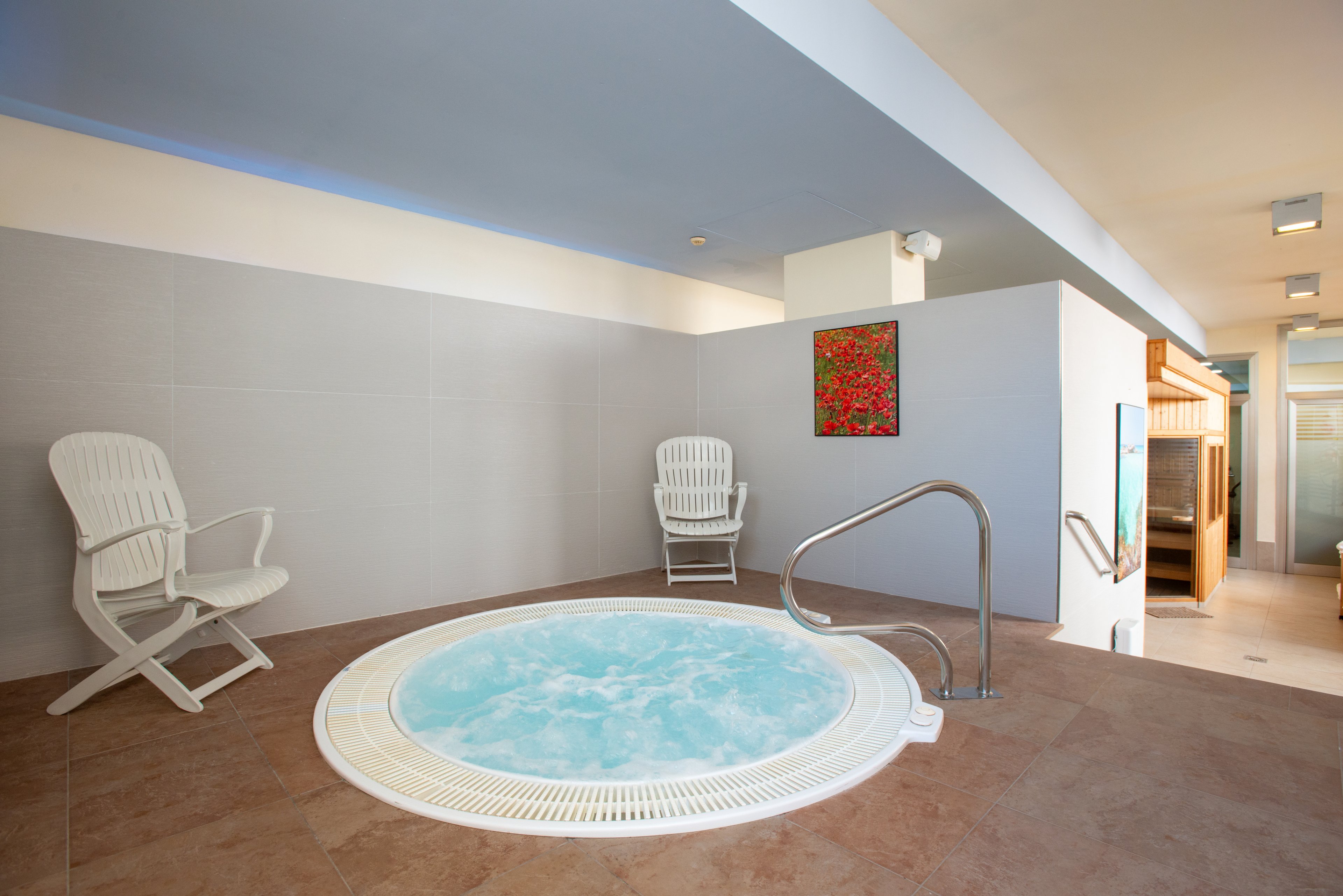 An indoor spa area features a round jacuzzi filled with bubbling water. Two white plastic lounge chairs sit on either side of the hot tub against a light gray wall. A colorful abstract painting hangs above the jacuzzi, adding a bright accent to the neutral space. Warm-toned floor tiles and stainless steel handrails complete the clean, modern wellness setting.