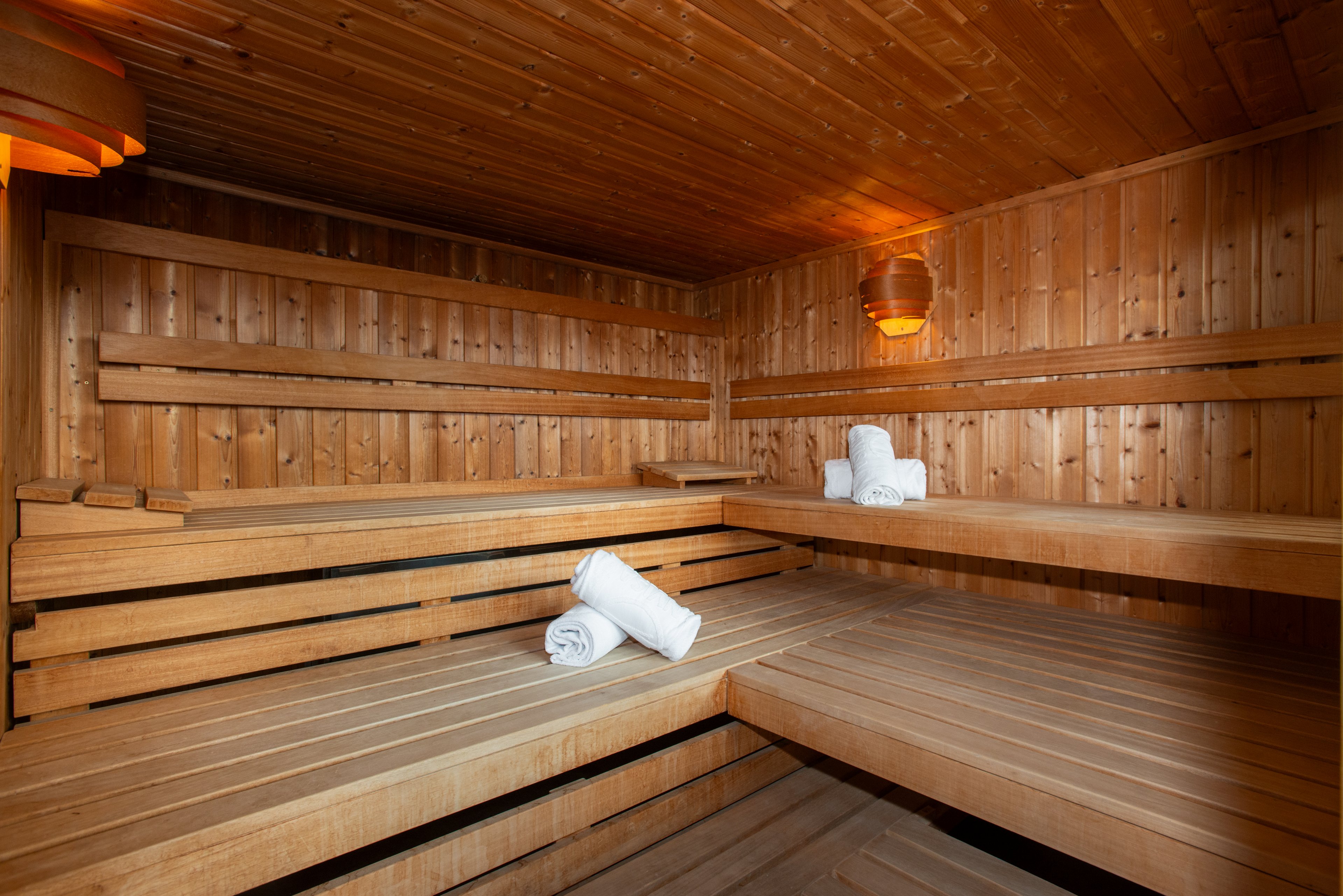 A spacious wooden sauna interior features tiered benches and warm ambient lighting. Rolled white towels are neatly placed on the benches, suggesting a prepared spa environment. The walls, ceiling, and seating are all made of smooth wood planks, creating a cohesive natural look. The scene is calm and inviting, emphasizing relaxation and wellness.