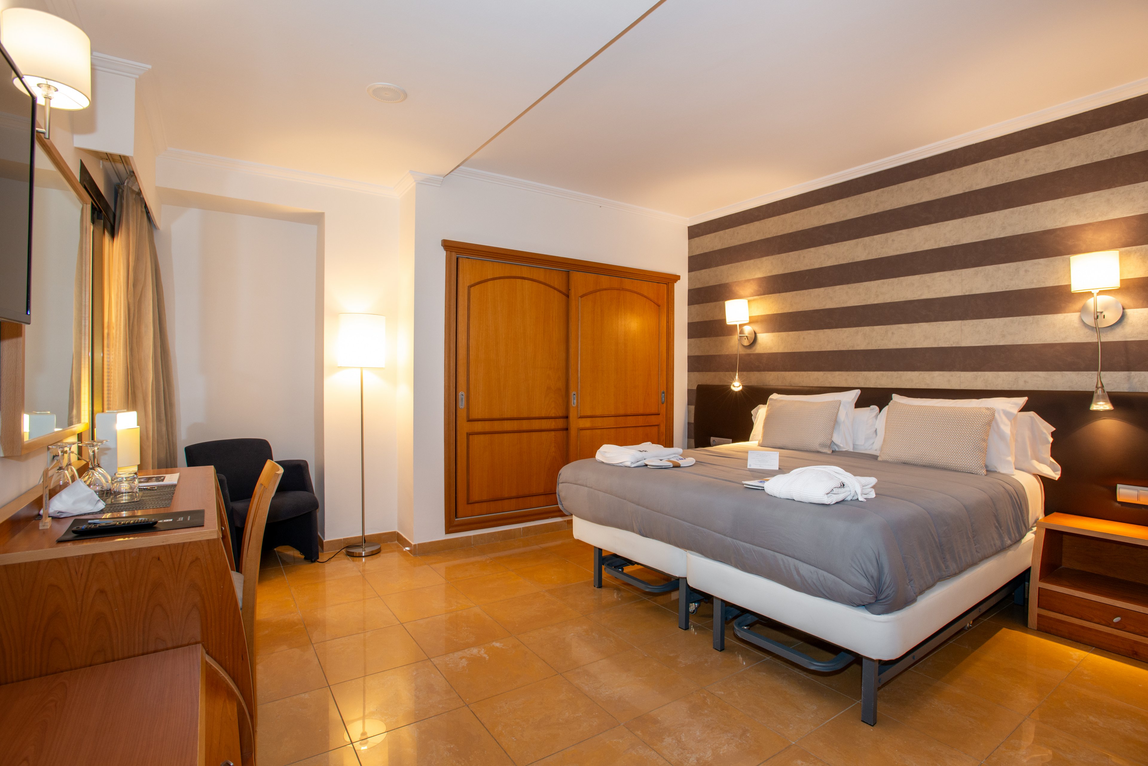 Well-lit hotel room featuring a large double bed with gray bedding and white pillows. The space includes wooden furniture, such as a wardrobe, bedside tables, and a desk with a mirror. Warm wall lamps and striped accent wall create a contemporary, inviting atmosphere. A small seating area with an armchair is visible near the window with curtains.