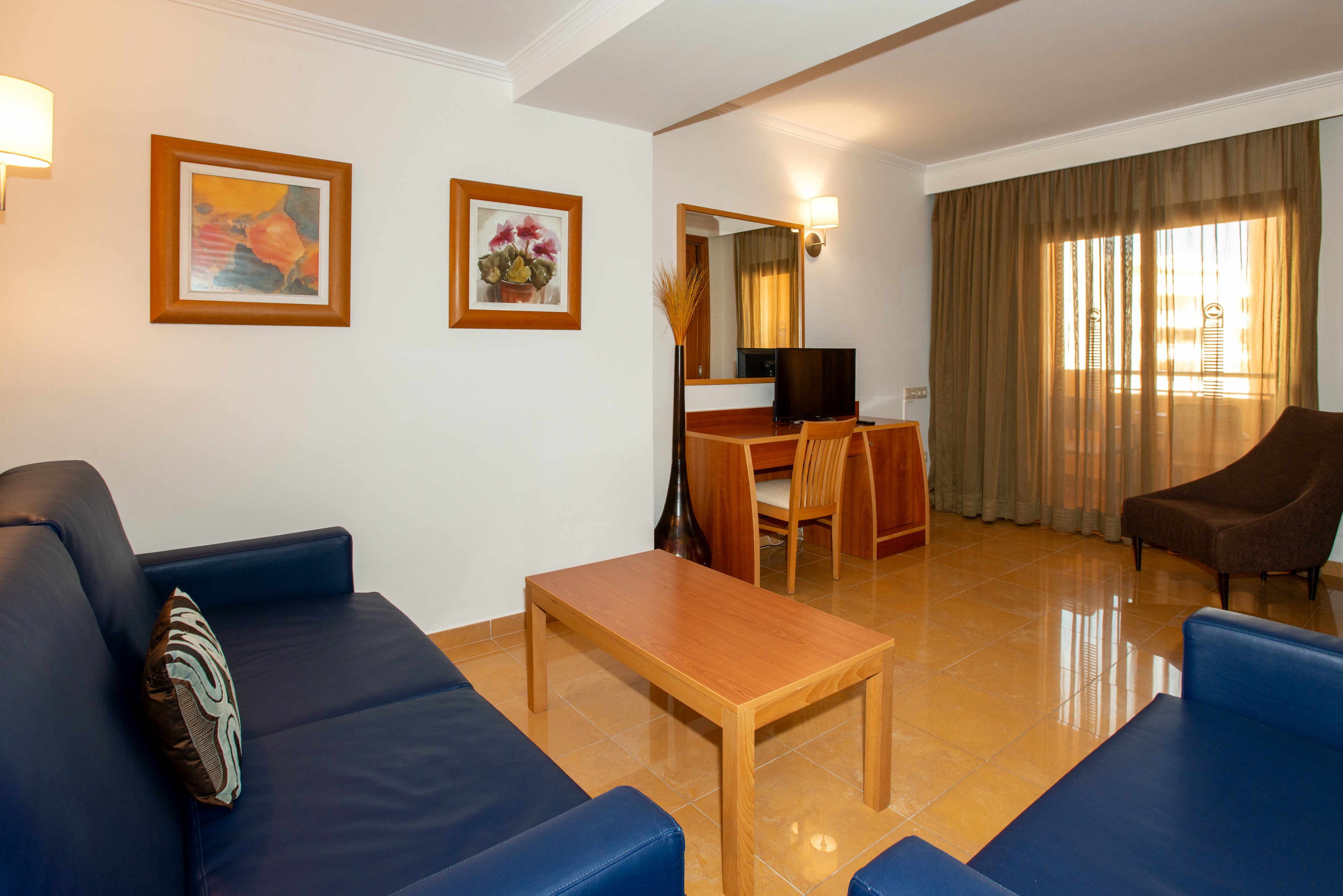 A tidy hotel living room features two blue leather sofas facing a simple wooden coffee table. In the background, a small desk with a chair and flat-screen television sits against the wall. Warm wall sconces and framed artwork add color to the neutral walls. Large curtains cover a sunlit window, creating a cozy yet spacious atmosphere.