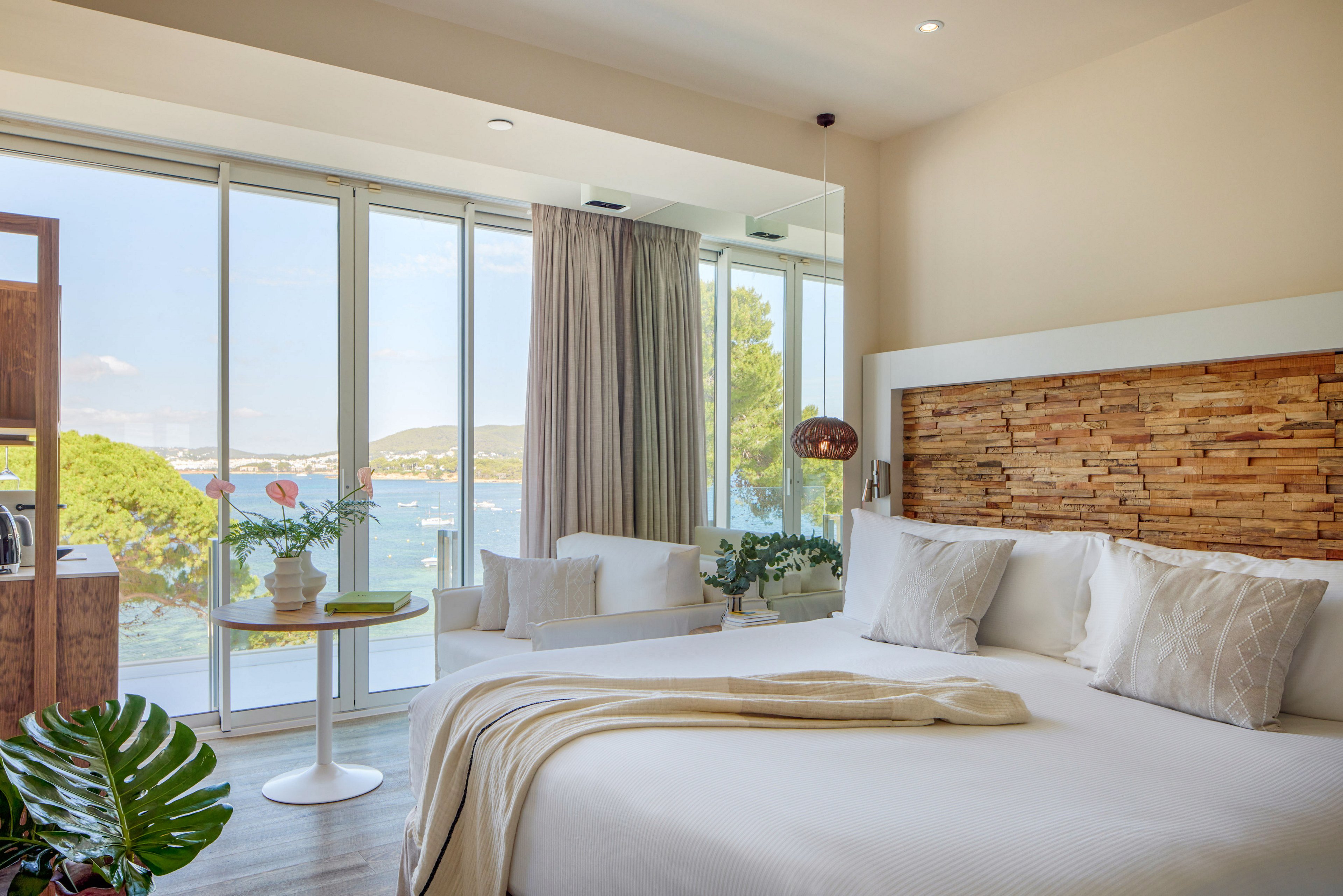 Contemporary hotel bedroom with a large bed faces floor-to-ceiling windows overlooking a scenic coastline. Natural light fills the room, highlighting neutral textiles, wooden accents, and indoor plants. A small seating area and side table sit near the window, offering a relaxing spot to enjoy the ocean view. The overall atmosphere is calm, airy, and inviting, ideal for a beachside retreat.