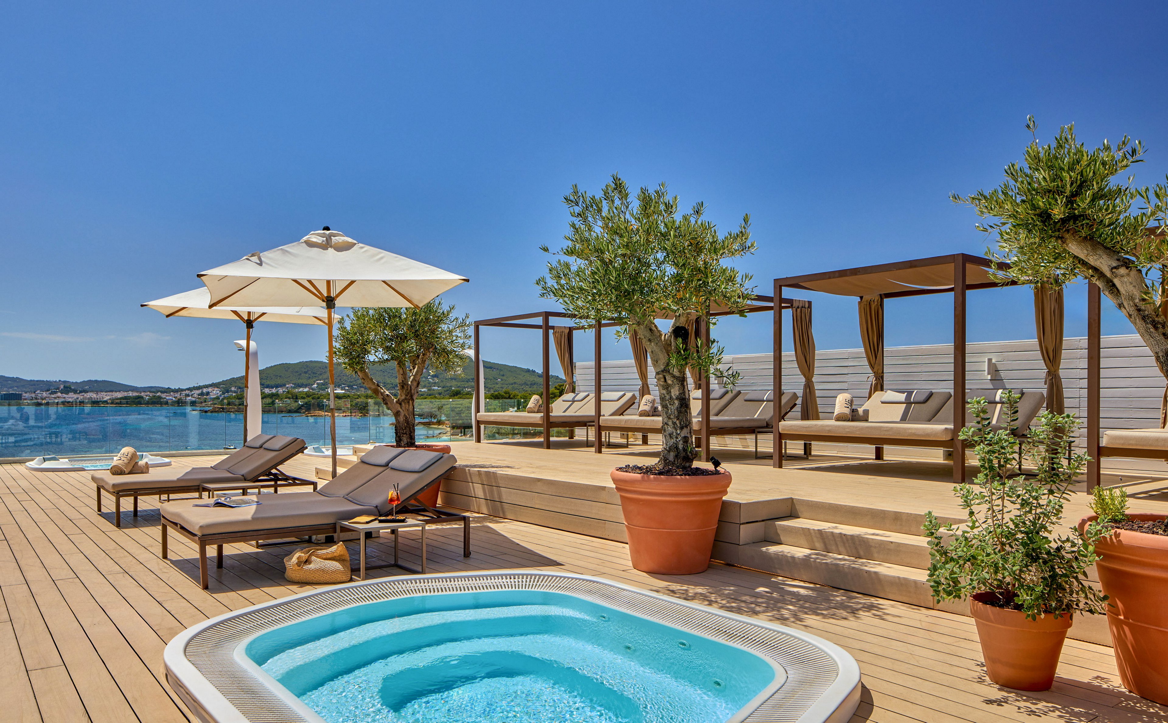 An upscale outdoor relaxation area features a small whirlpool pool set into a wooden deck overlooking the sea. Sun loungers with cushions and white umbrellas are arranged nearby, with cabana-style daybeds in the background. Large potted trees and plants add greenery to the warm, minimalist design. Calm blue water and distant hills complete the serene coastal setting under a clear sky.