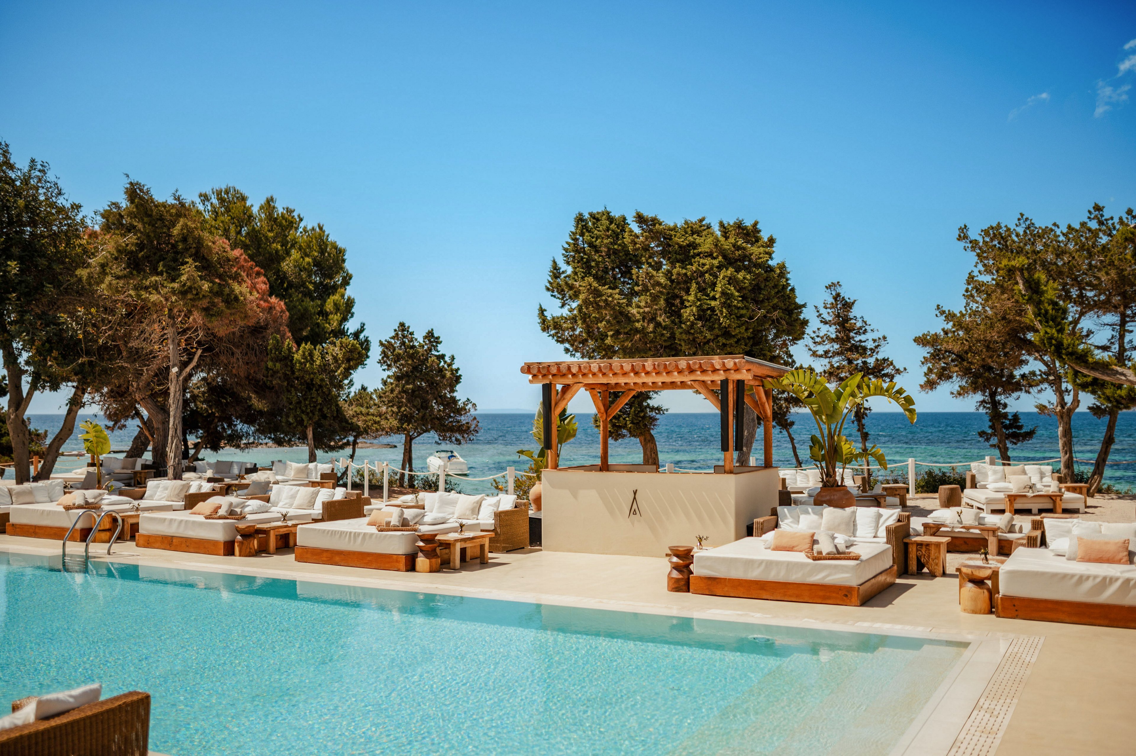An upscale outdoor pool area overlooks a calm blue sea under a clear sky. Wooden cabanas and cushioned sunbeds surround the turquoise swimming pool, creating a relaxed resort atmosphere. Tall trees and tropical plants frame the scene, adding natural shade and texture. The setting suggests a tranquil beach club or coastal hotel designed for leisure and sunbathing.