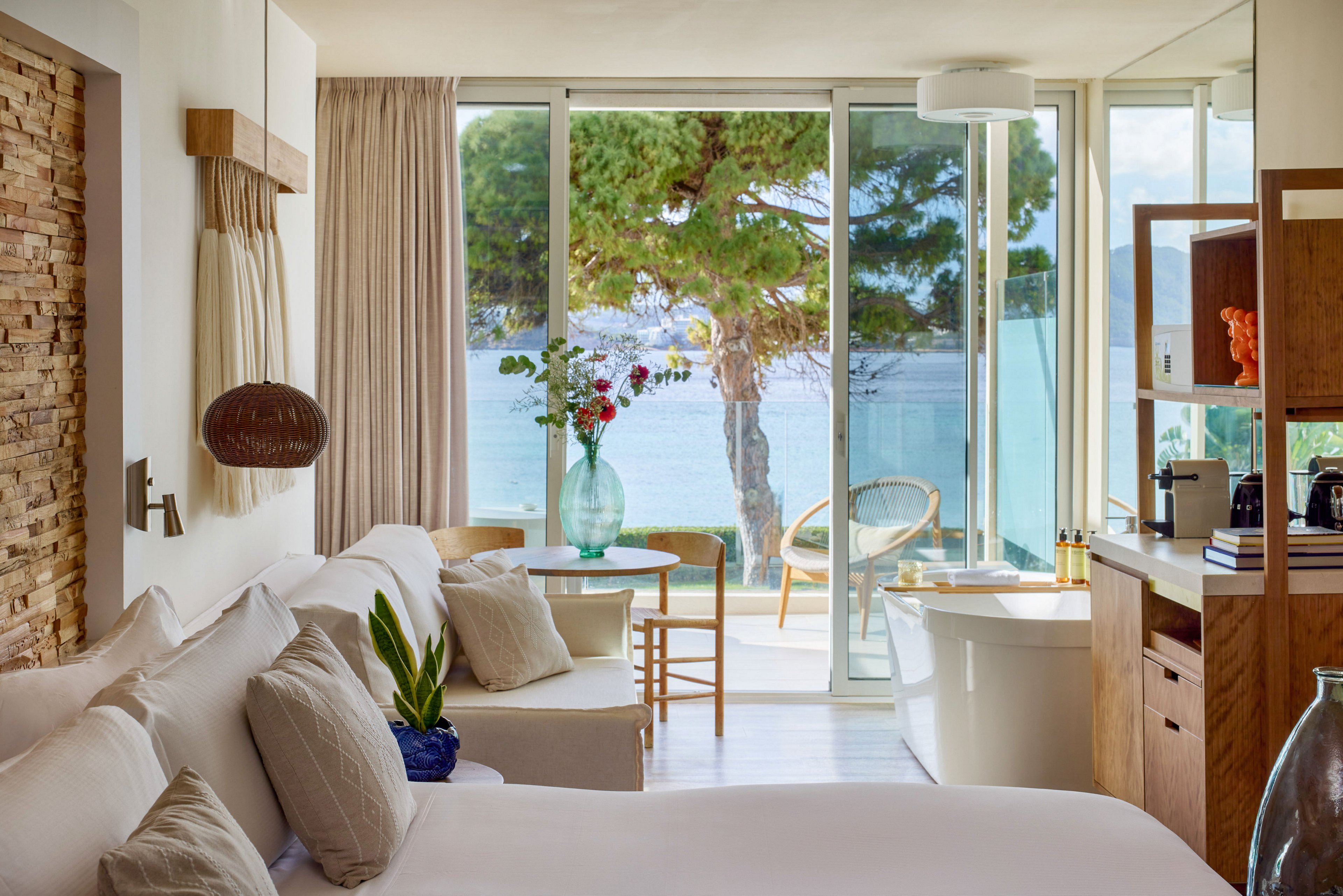 A contemporary hotel room features a large bed and soft neutral furnishings. Floor-to-ceiling glass doors open onto a balcony overlooking the sea and pine trees. A small seating area with a round wooden table, chairs, and a glass vase of flowers sits near the window. Warm wood accents, stone wall details, and natural light create a calm, inviting atmosphere.