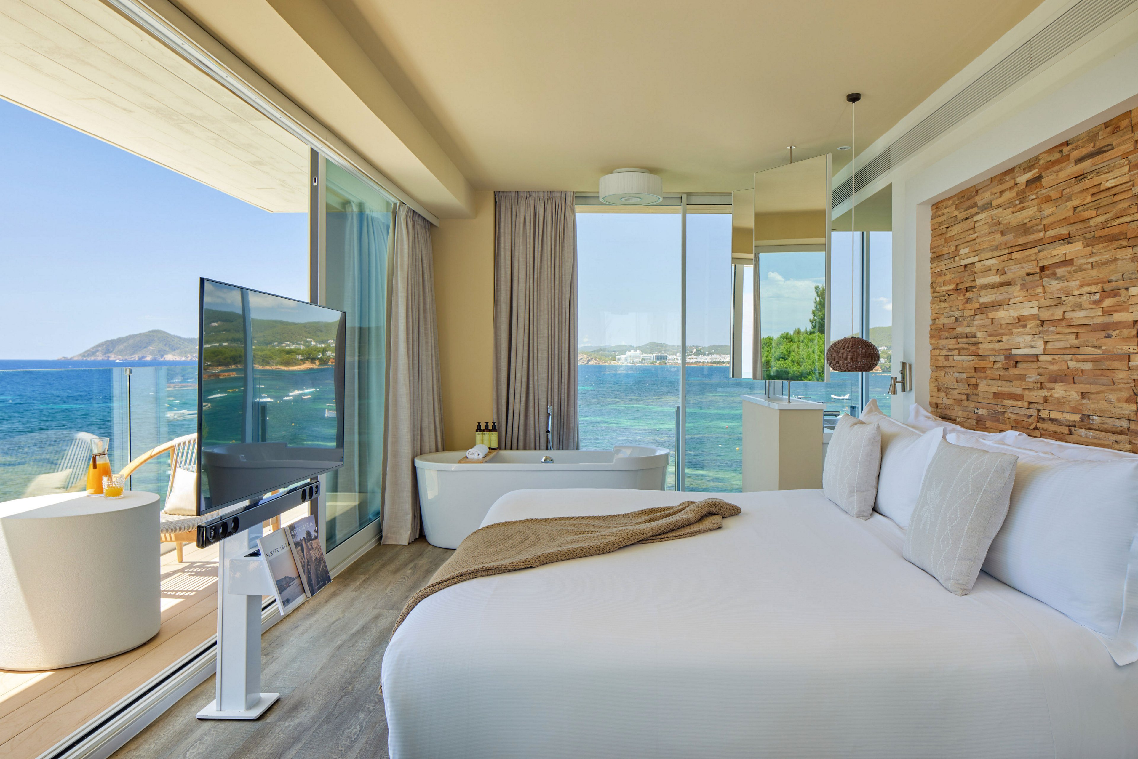 Bright contemporary hotel room features a large bed facing a flat-screen TV and floor-to-ceiling windows. A freestanding bathtub sits near the glass wall, opening onto a private balcony with seating and panoramic sea views. Natural wood textures on the accent wall and neutral furnishings create a calm, minimalist atmosphere. Sunlit blue water and distant coastline enhance the luxurious vacation setting.