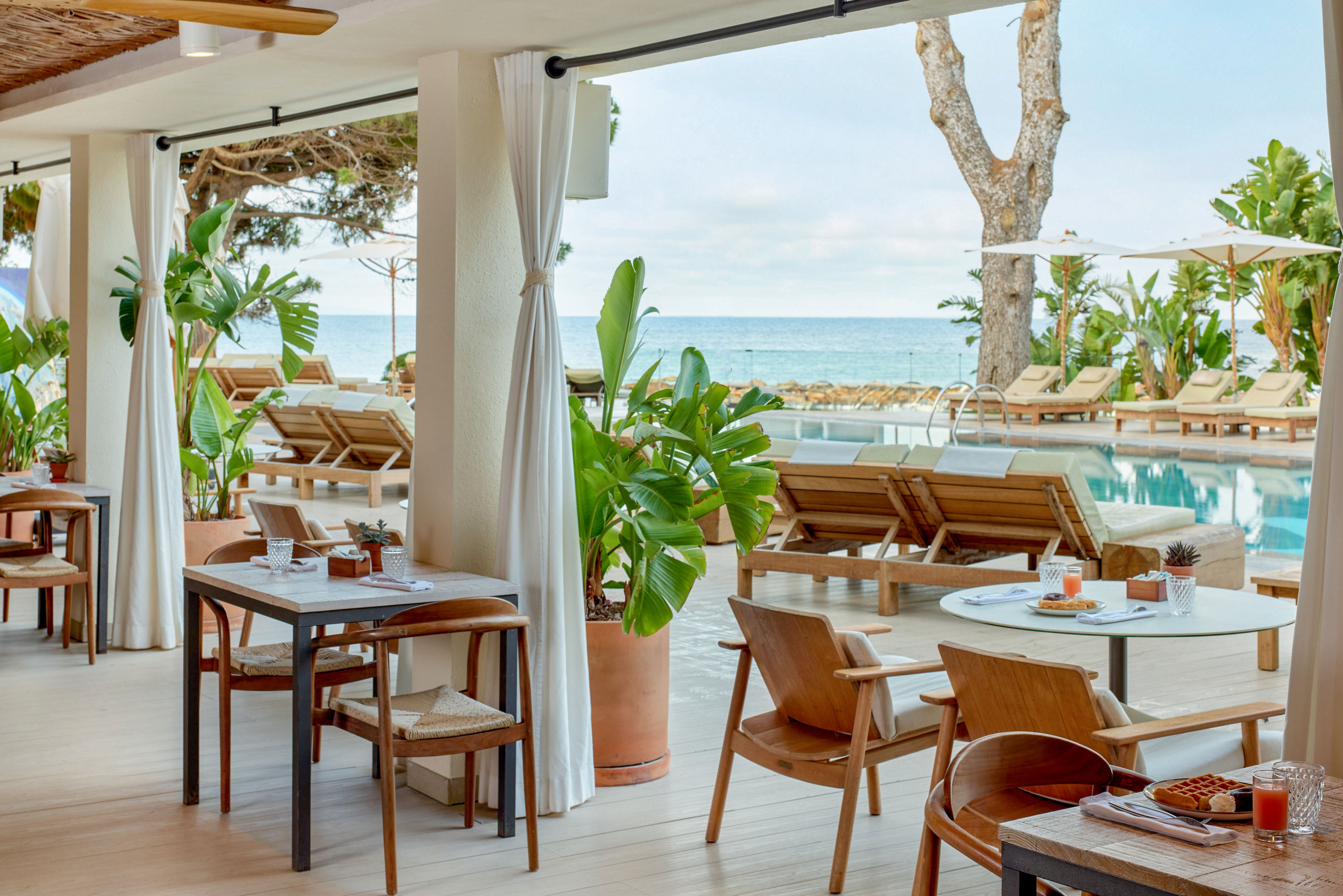 An open-air resort terrace overlooks a swimming pool and the ocean beyond. Wooden tables and chairs are neatly arranged with drinks and table settings, creating a relaxed dining atmosphere. Sun loungers line the poolside under large white umbrellas, surrounded by lush tropical plants. Soft natural light and neutral tones give the scene a calm, upscale vacation feel.
