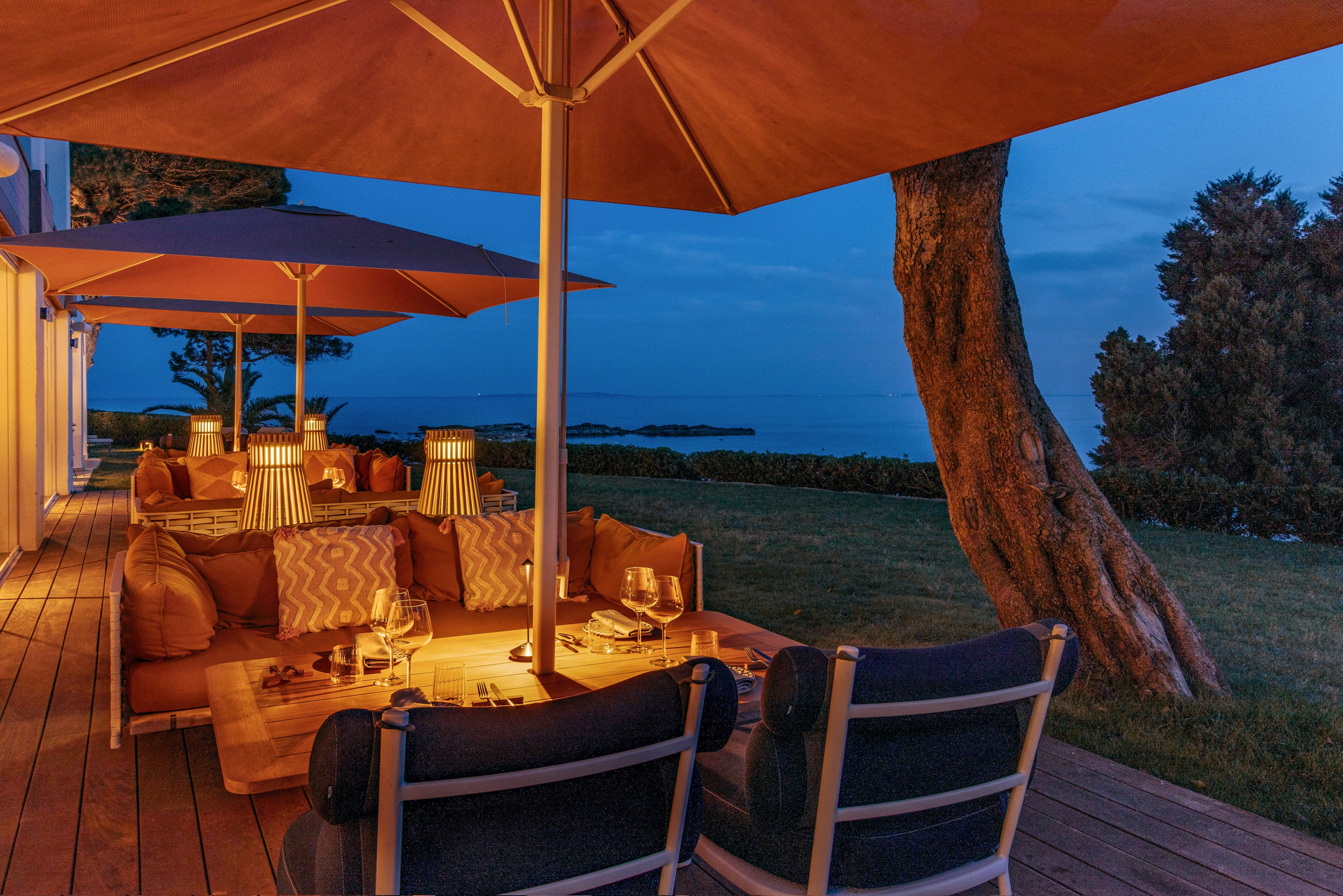 An inviting outdoor lounge terrace is set under large umbrellas, softly lit by warm table lamps and candles. Cushioned sofas and armchairs surround low wooden tables with wine glasses and tableware, creating an intimate dining atmosphere. The scene overlooks a calm sea and distant islands at blue hour, with a large tree framing the view. The combination of warm interior light and cool evening sky evokes a relaxed, romantic mood.