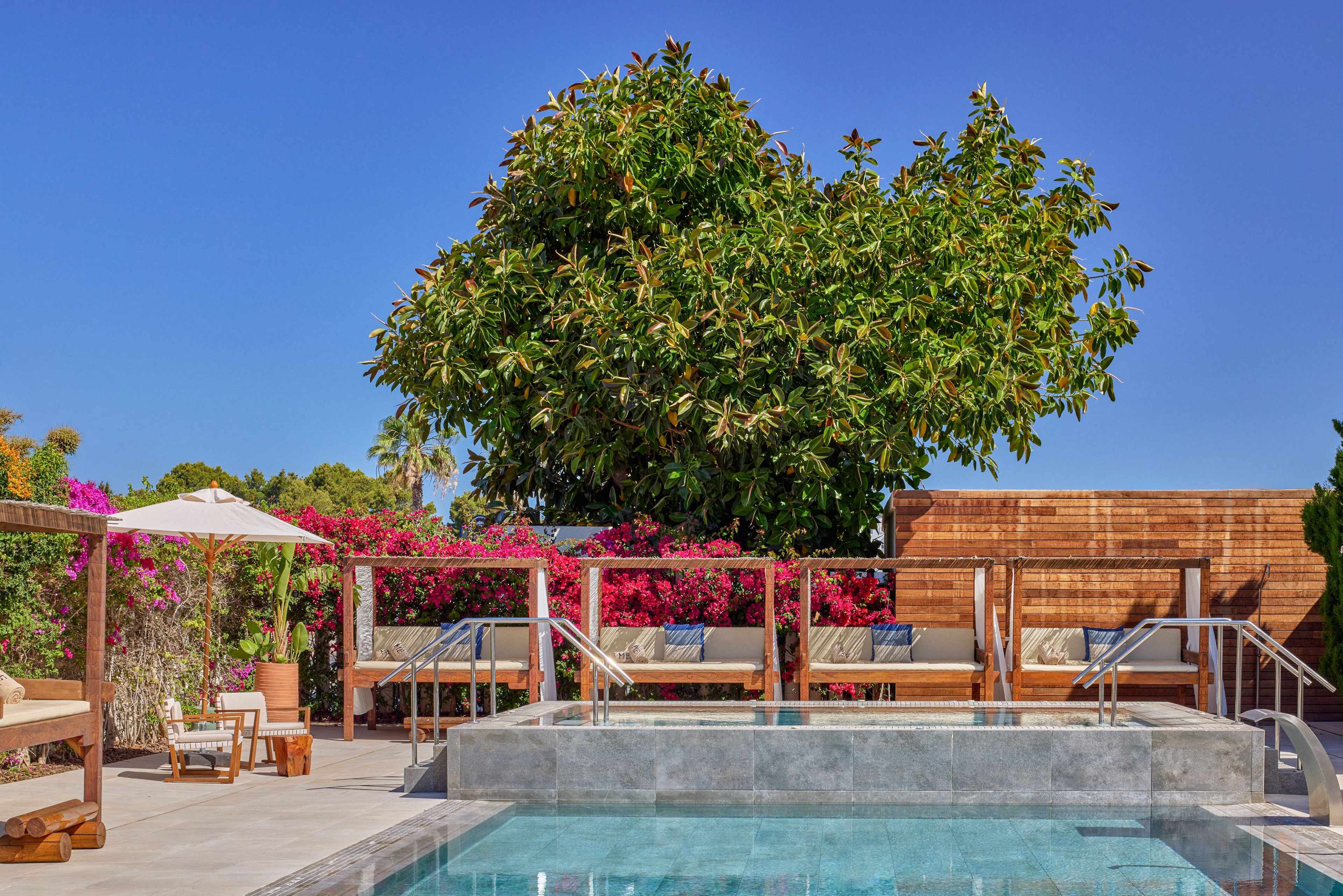 A contemporary outdoor swimming pool is surrounded by sleek stone decking and wooden cabanas with cushioned seating. Bright pink flowering shrubs and lush green trees frame the space, adding vibrant color against the clear blue sky. Metal handrails lead into the pool, while sun loungers and a white umbrella provide shaded relaxation areas. The scene suggests a tranquil resort or luxury backyard designed for leisure and comfort.