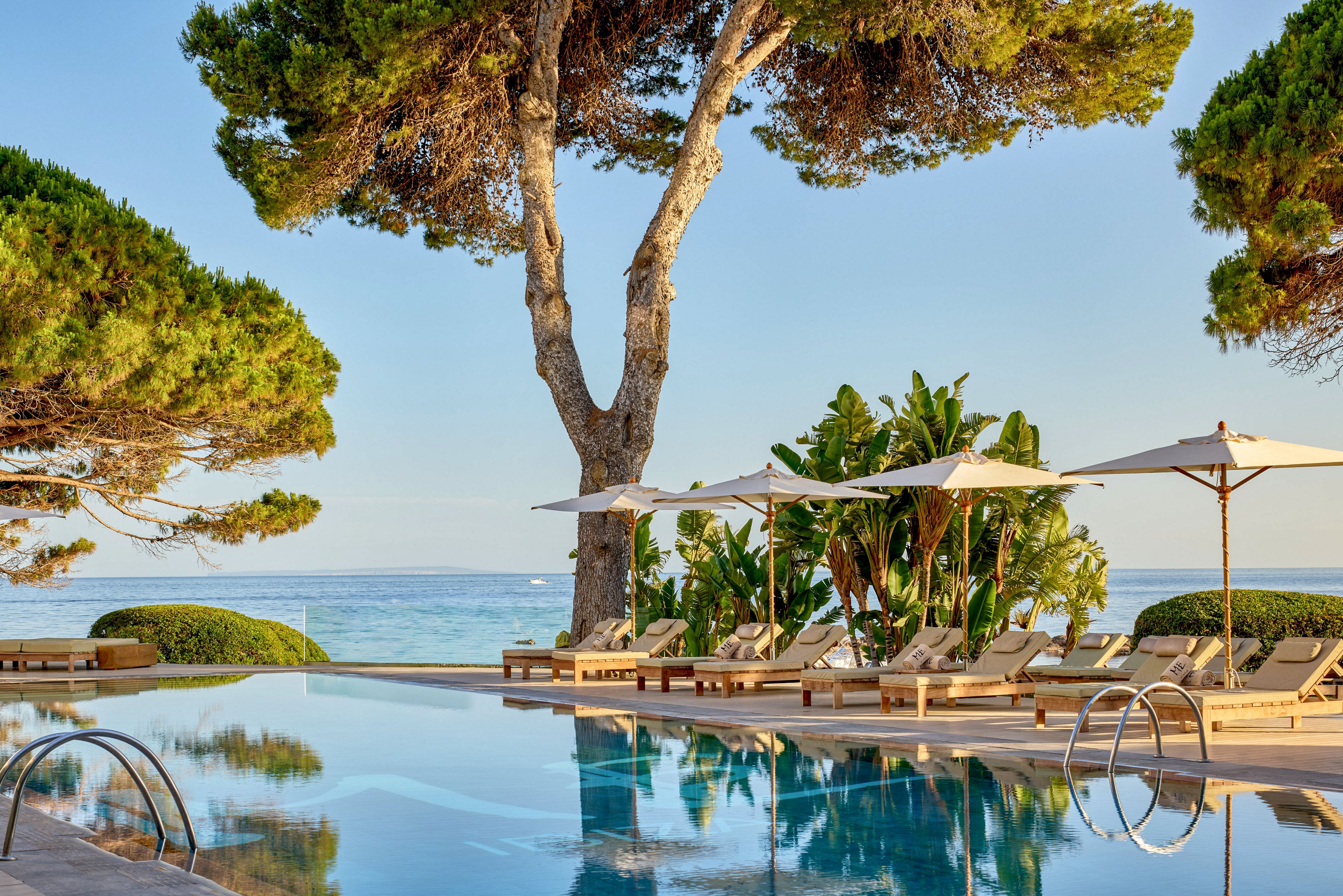 A tranquil outdoor swimming pool sits beside a row of cushioned sun loungers under large white umbrellas. Tall trees and lush tropical plants frame the scene, leading the eye toward the calm blue sea in the background. The clear water of the pool reflects the surrounding greenery and umbrellas, enhancing the serene atmosphere. Warm sunlight suggests a peaceful vacation setting on a coastal resort.