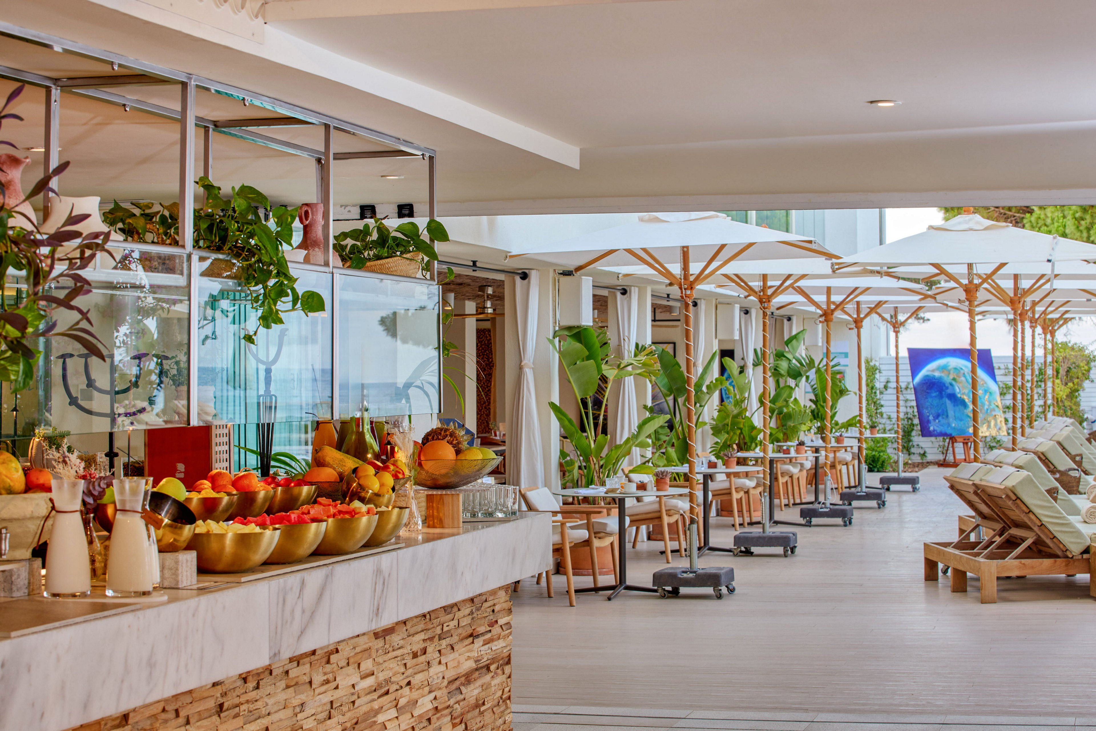 An open-air resort bar displays bowls of fresh fruit and drink ingredients on a marble counter with a stone base. The bar faces a long terrace lined with wooden tables, chairs, and large white umbrellas. Green potted plants add a tropical feel along the walkway, leading toward sun loungers arranged in rows. Bright, natural daylight and warm tones create a relaxed vacation atmosphere by the water.