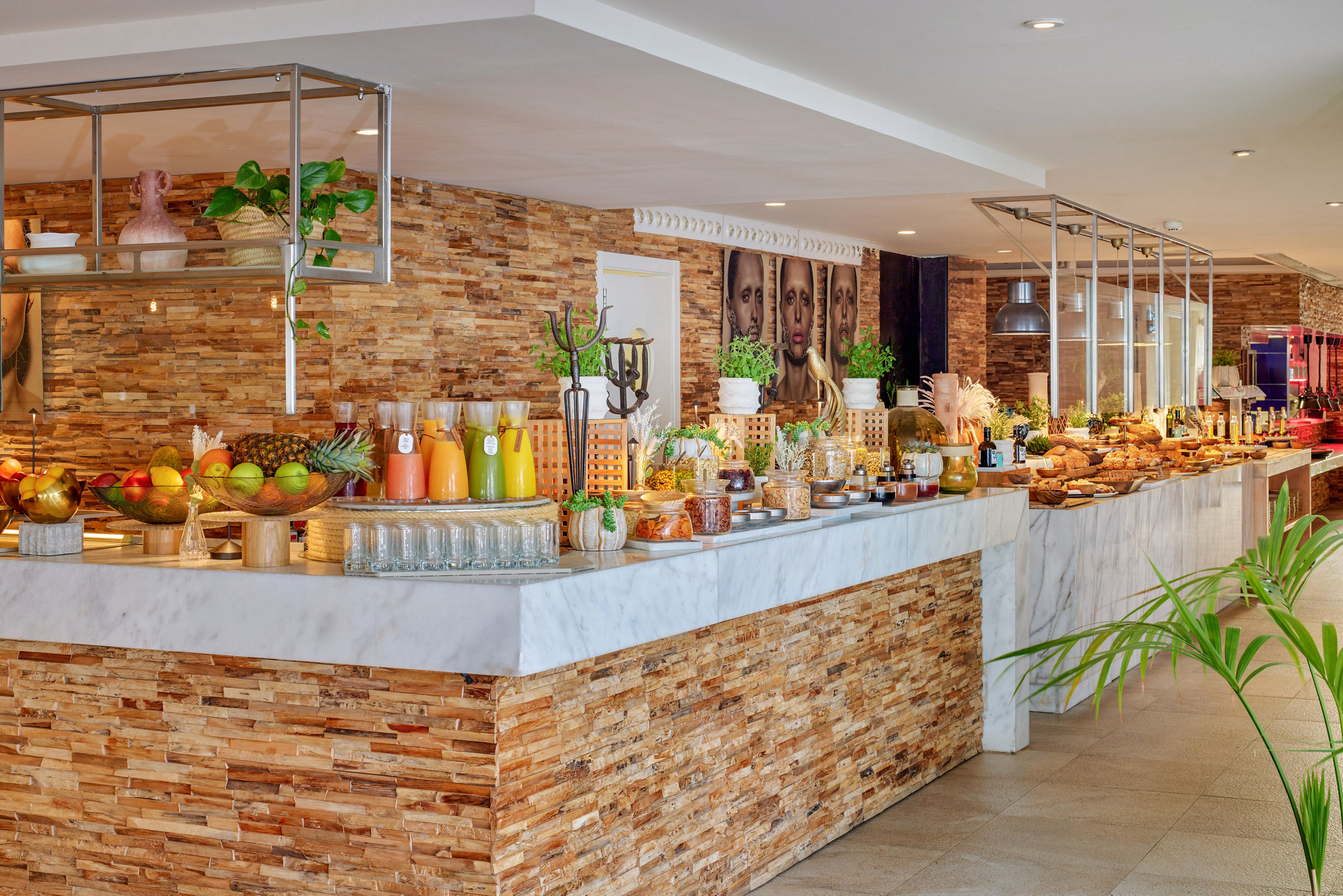 A spacious hotel buffet counter is filled with assorted breakfast foods and colorful beverages. Glass dispensers and bottles of juice are arranged alongside fresh fruits and pastries on a marble and stone counter. Warm lighting and decorative plants create an inviting, contemporary dining atmosphere. The setting suggests a self-service breakfast area in a modern hotel or resort.
